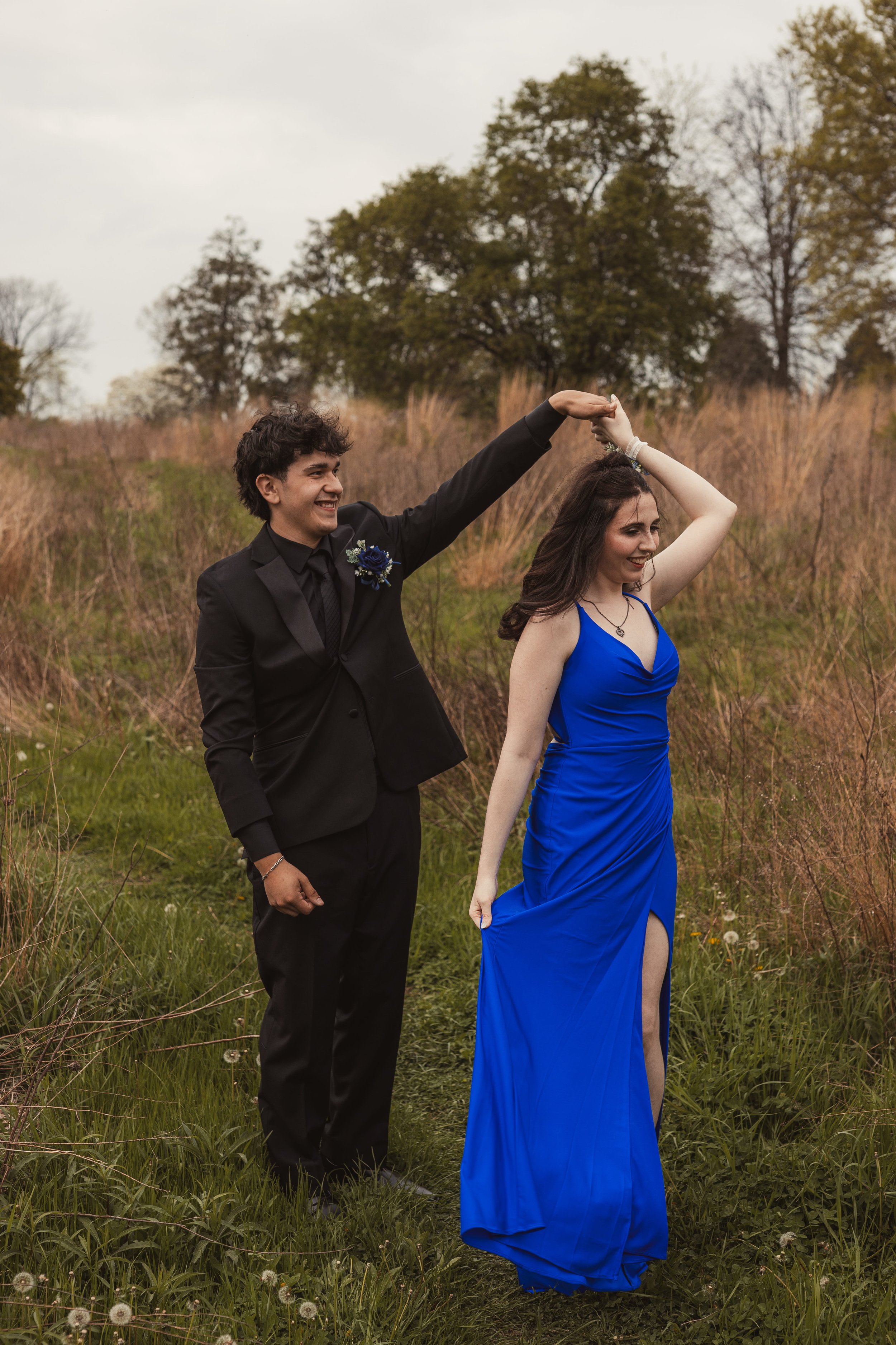 A young man and woman dancing outdoors in a field. The man is wearing a suit with a boutonniere, and the woman is wearing a blue gown. The man is holding the woman's hand up as they dance. Trees are visible in the background on an overcast day.