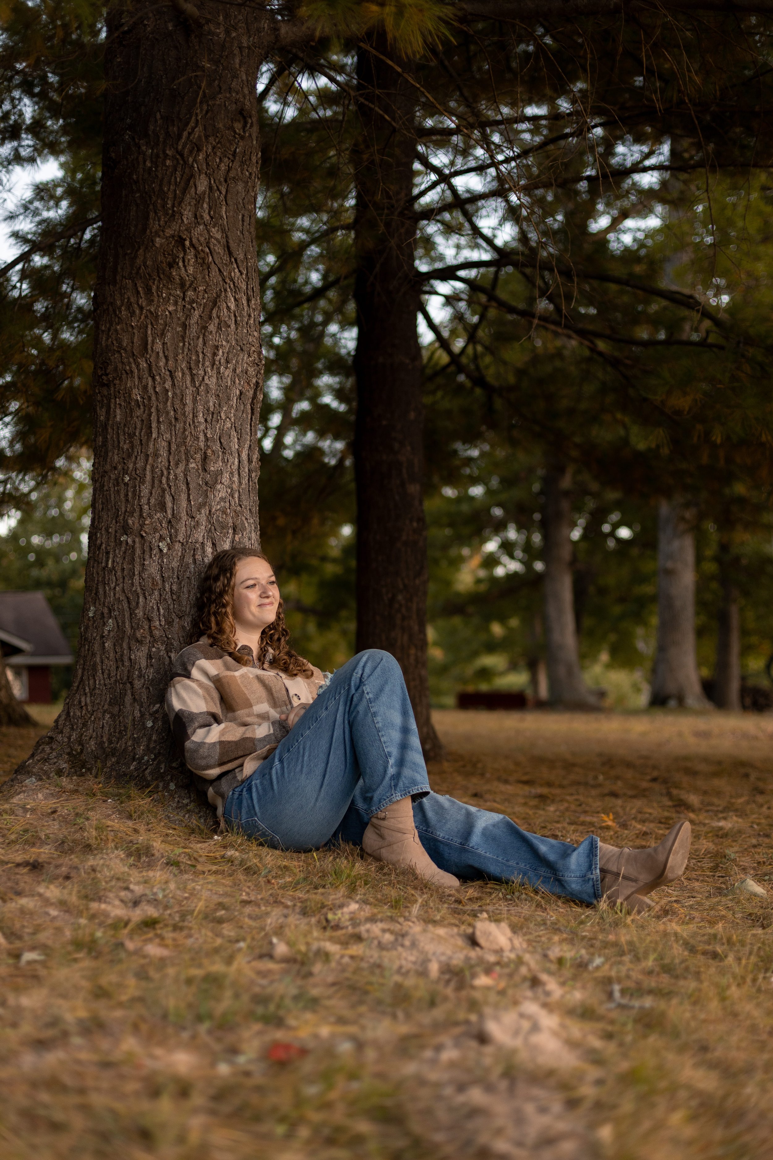 Young woman in jeans and plaid flannel sitting against a pine tree in a moody fall forest, senior portrait