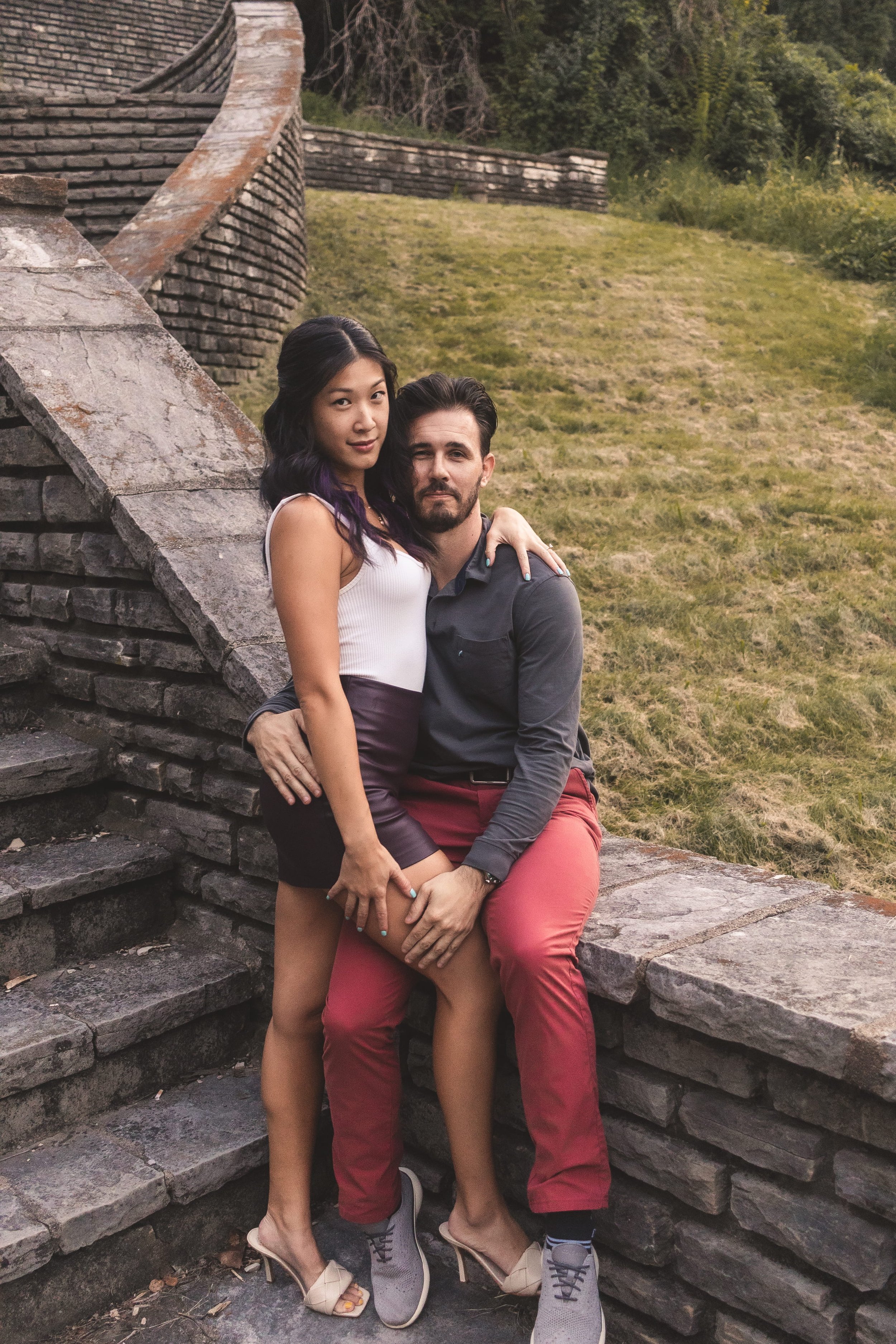 A young woman and a man sitting on stone stairs outdoors, with a grassy hill and trees in the background. The woman has long dark hair with purple streaks, wearing a white tank top, dark shorts, and high heels. The man has dark hair and a beard, wear