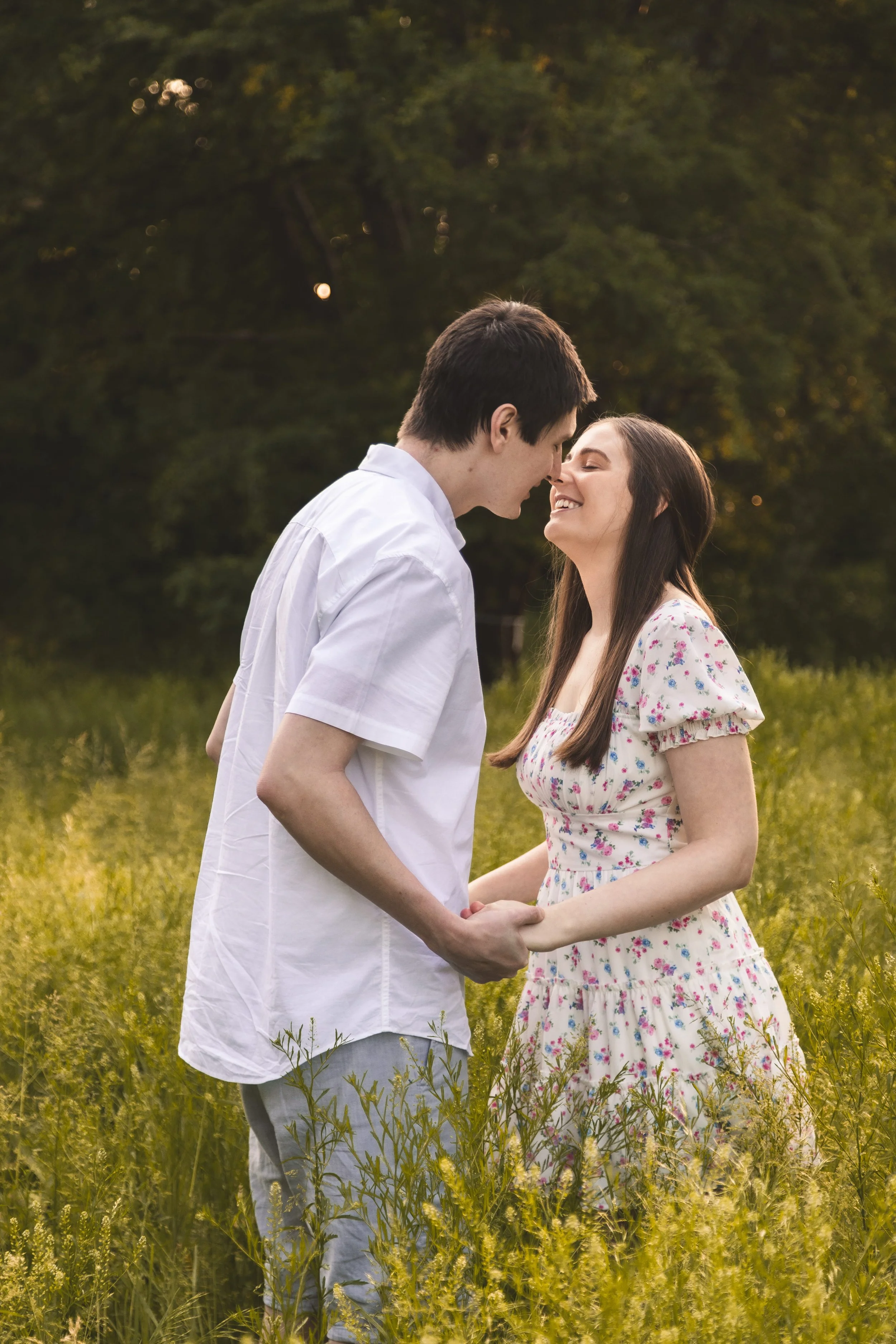A young couple with light skin holding hands and leaning in to touch noses in a green field during golden hour, surrounded by tall grass and trees.