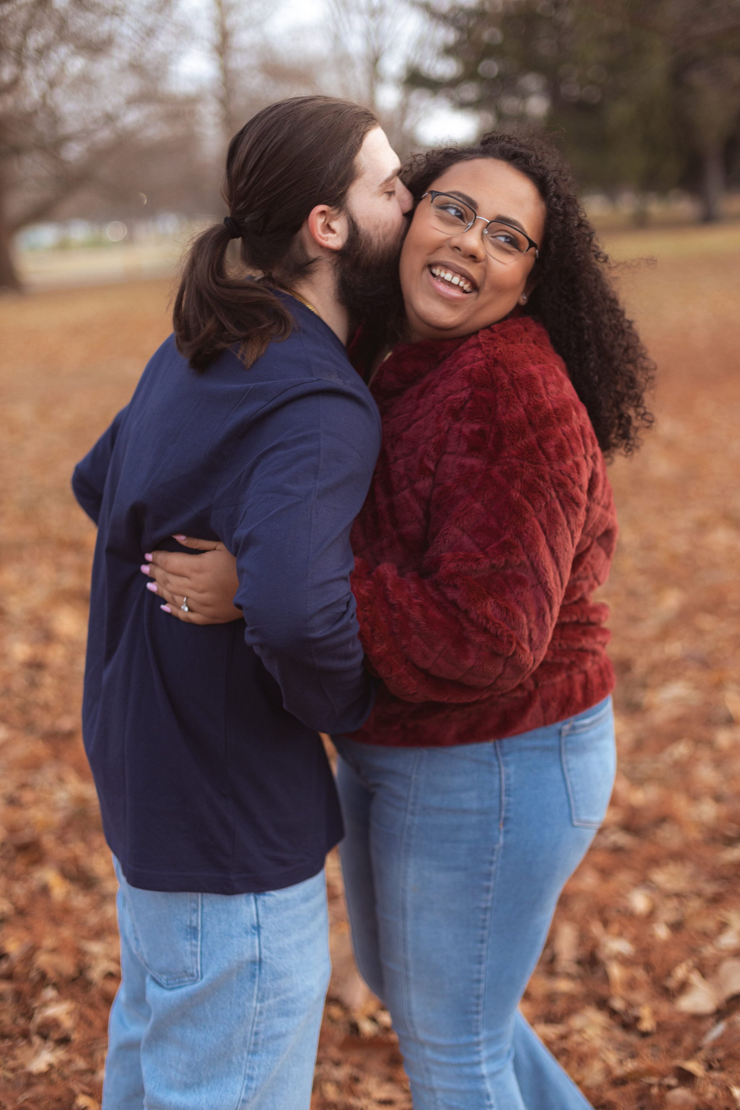 Couple in a fall park, man in navy hoodie kissing his partner on the cheek as she laughs