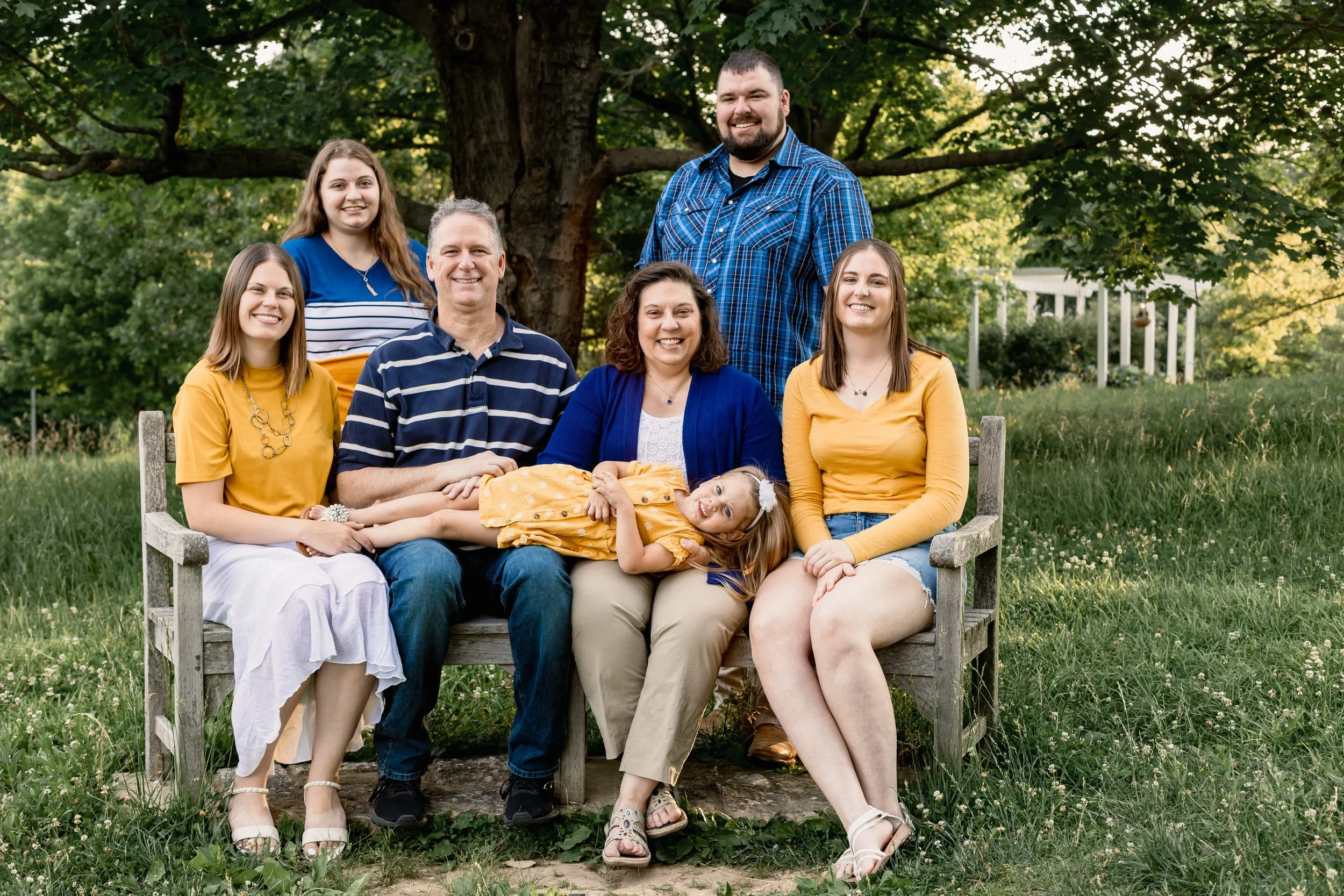 Family of seven posing on a bench under a large tree in coordinating yellow and blue outfits