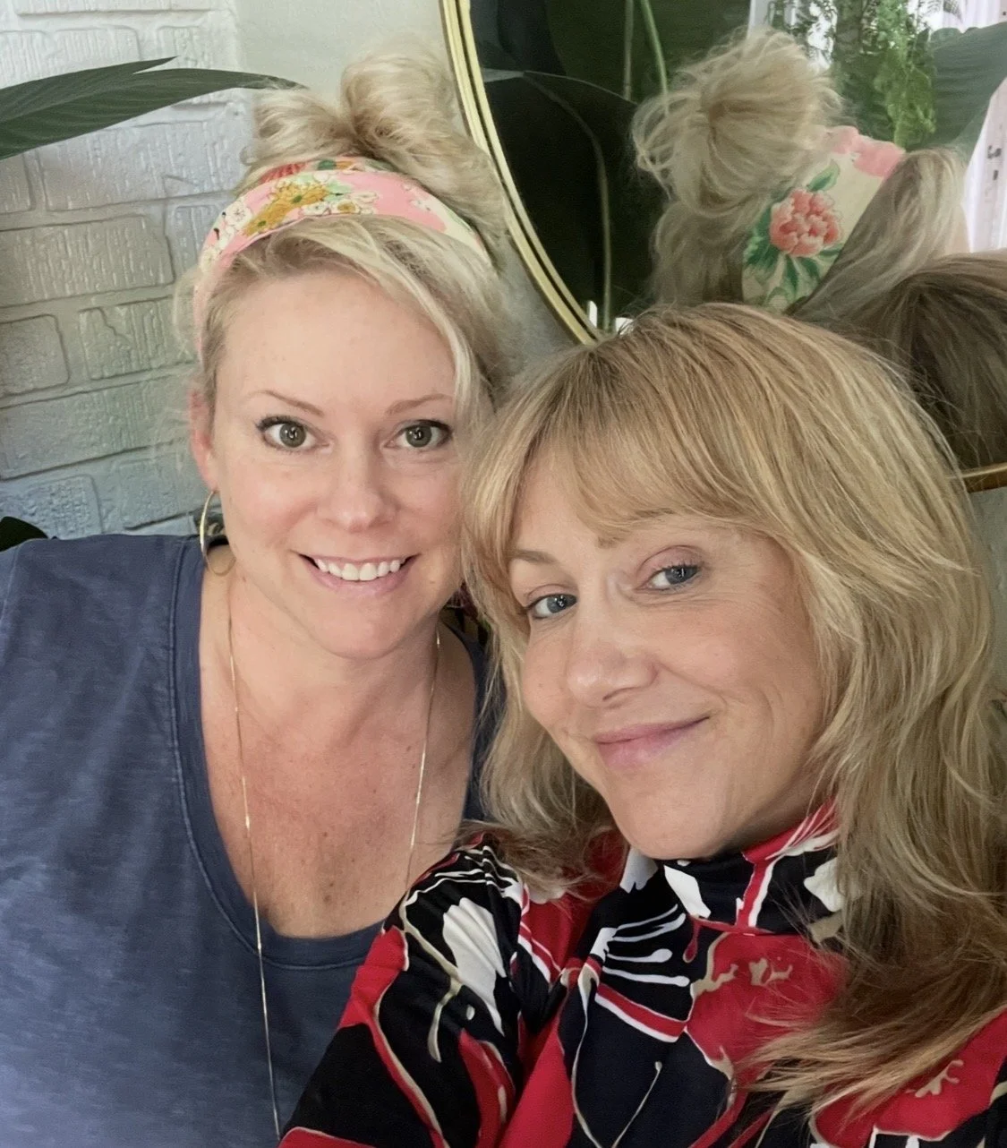 Two women smiling and taking a selfie in front of a mirror, with one woman wearing a pink floral headband and a dark T-shirt, and the other wearing a colorful patterned blouse. Mirrors and plants are visible in the background.