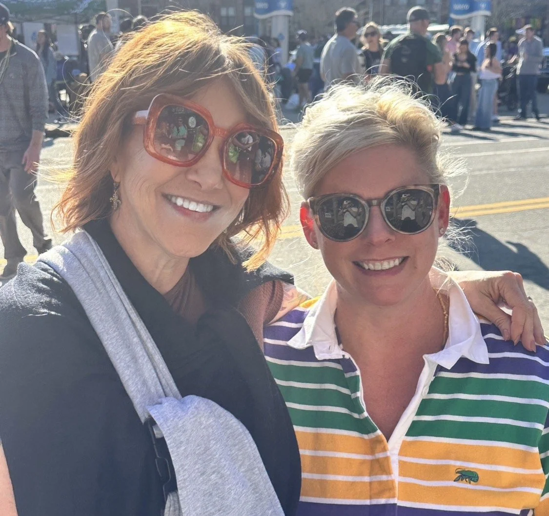 Two women smiling and posing for a photo outdoors in a busy urban setting, with other people walking and standing in the background.
