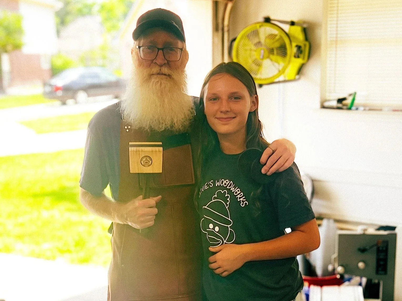 A volunteer woodworker poses with a woodworker with a disability in his garage workshop, surrounded by tools and equipment, after helping build custom adaptive woodworking hardware to support his continued craft.