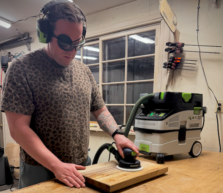 Woodworker using a Festool random orbital sander with dust extraction on a hardwood cutting board in a home workshop, wearing hearing and eye protection.