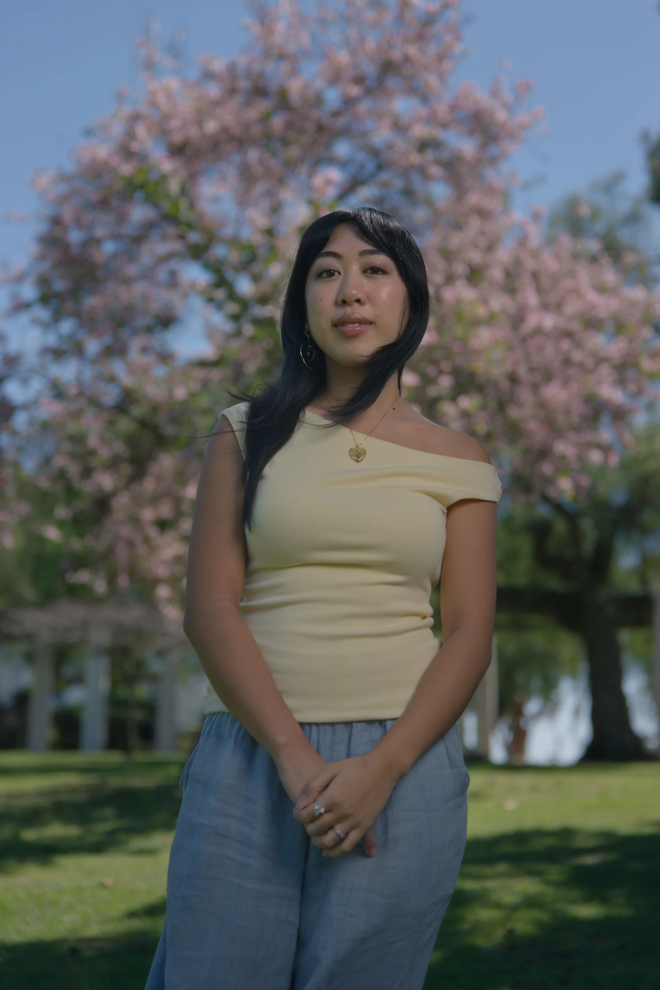 A woman with long black hair, wearing a yellow off-shoulder top and gray pants, stands outdoors in front of a pink flowering tree on a sunny day.