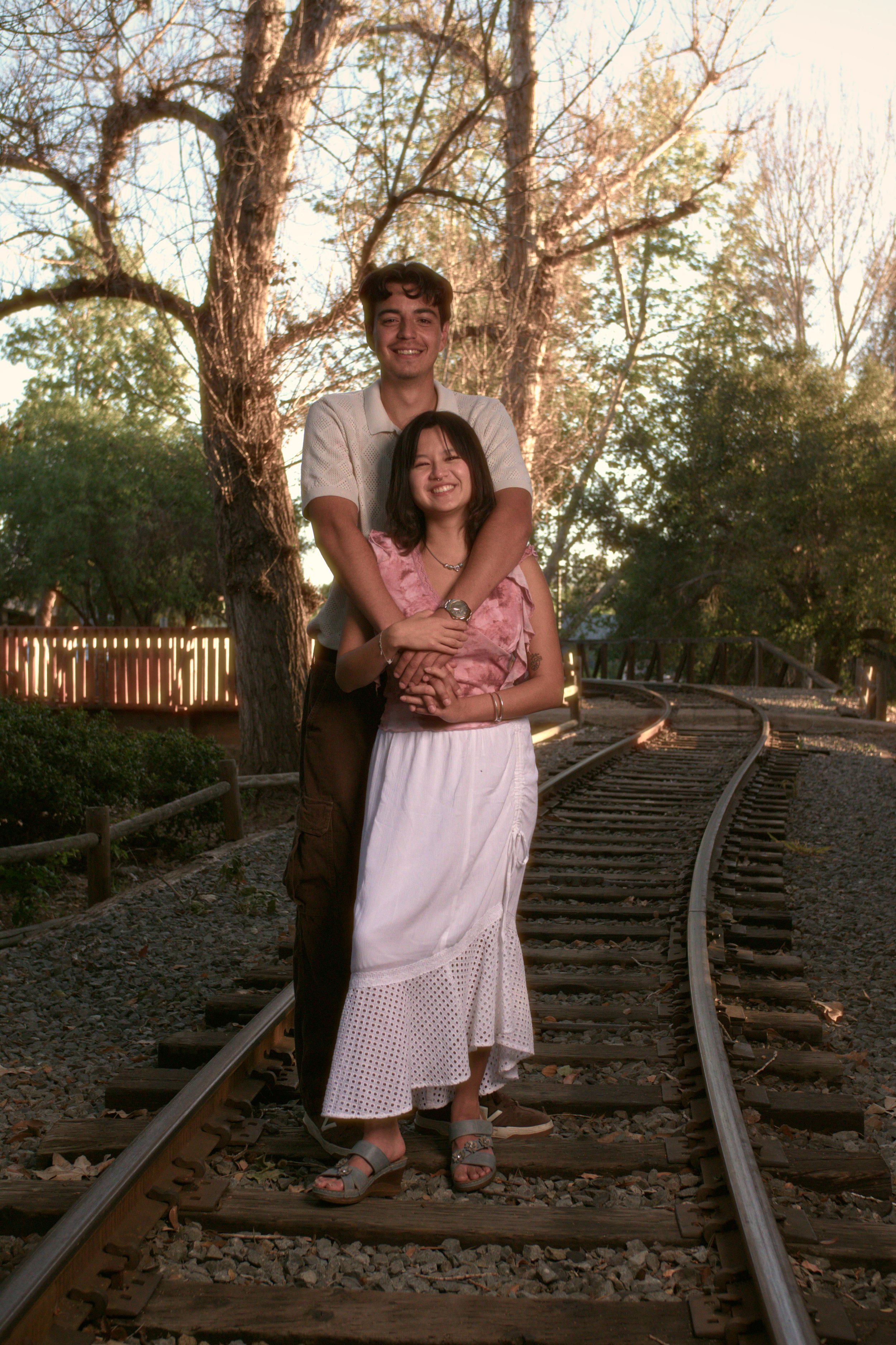A young man and woman standing together on a railroad track surrounded by trees and sunlight, smiling.