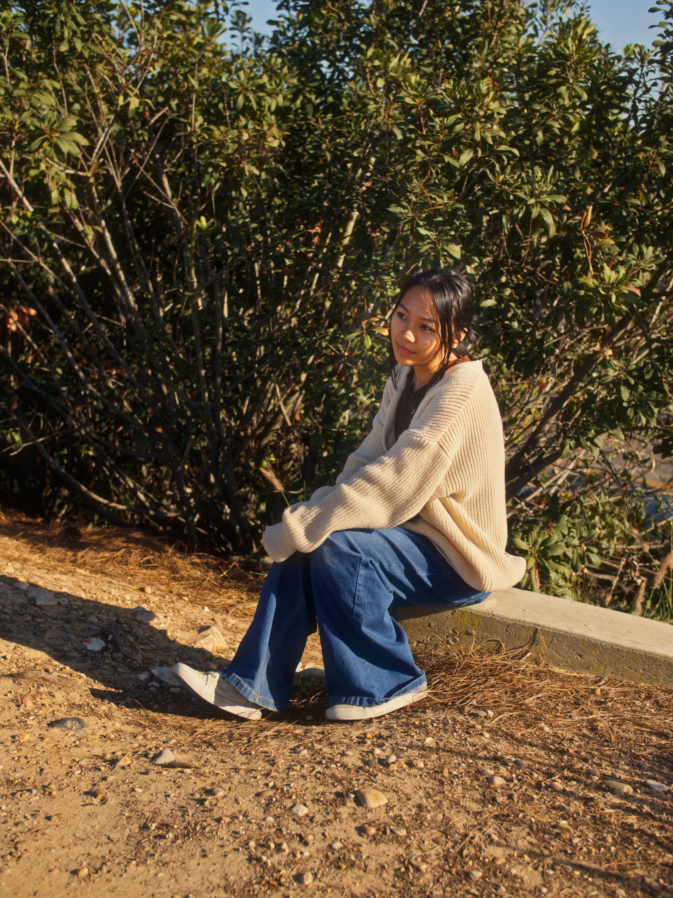 A young woman wearing a beige sweater with blue jeans and sneakers sits on a concrete ledge on a dirt path, surrounded by green bushes, during daylight.