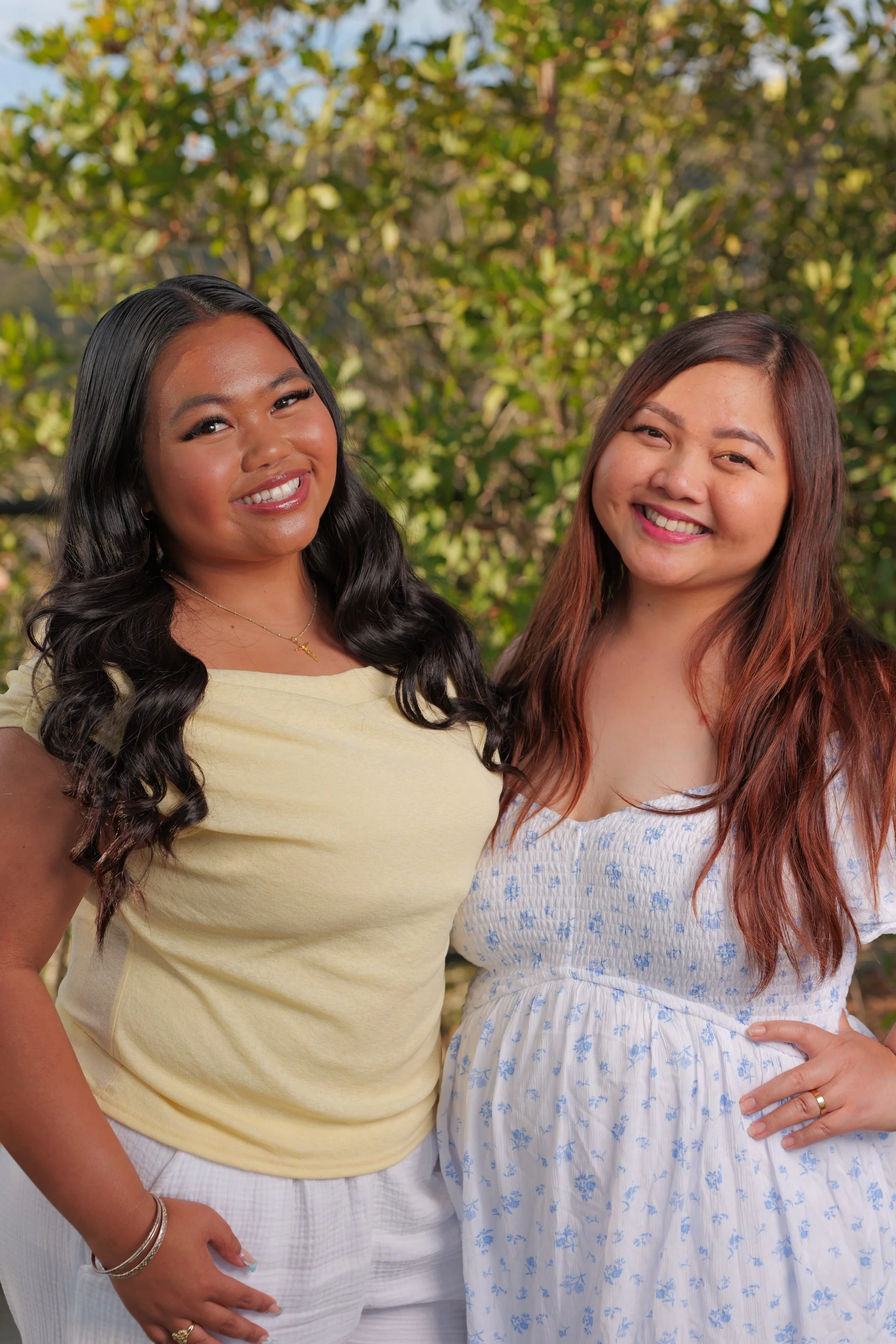Two women smiling outdoors in front of trees, one with long black hair and the other with long reddish-brown hair.