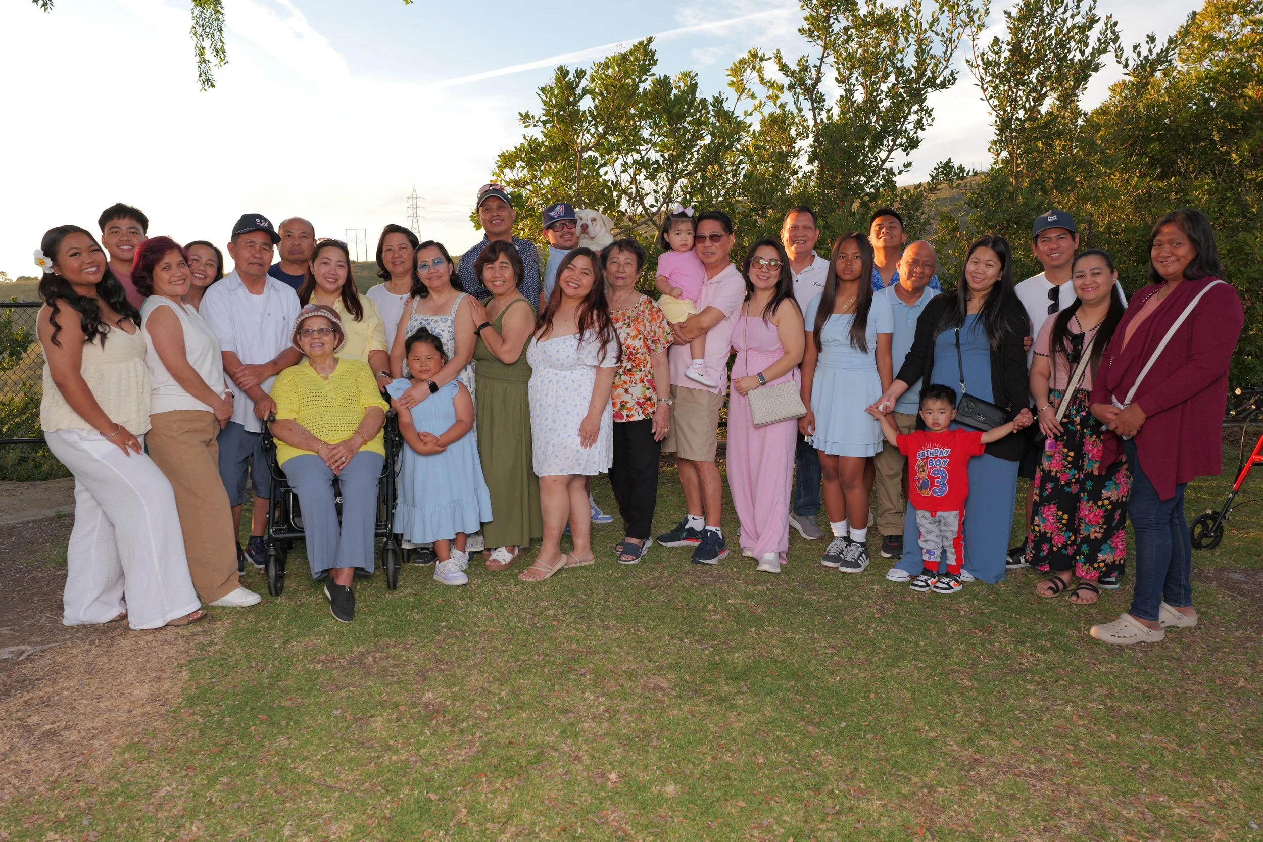 Group of people posing outdoors with trees and sky in the background, including elderly, adults, and children.