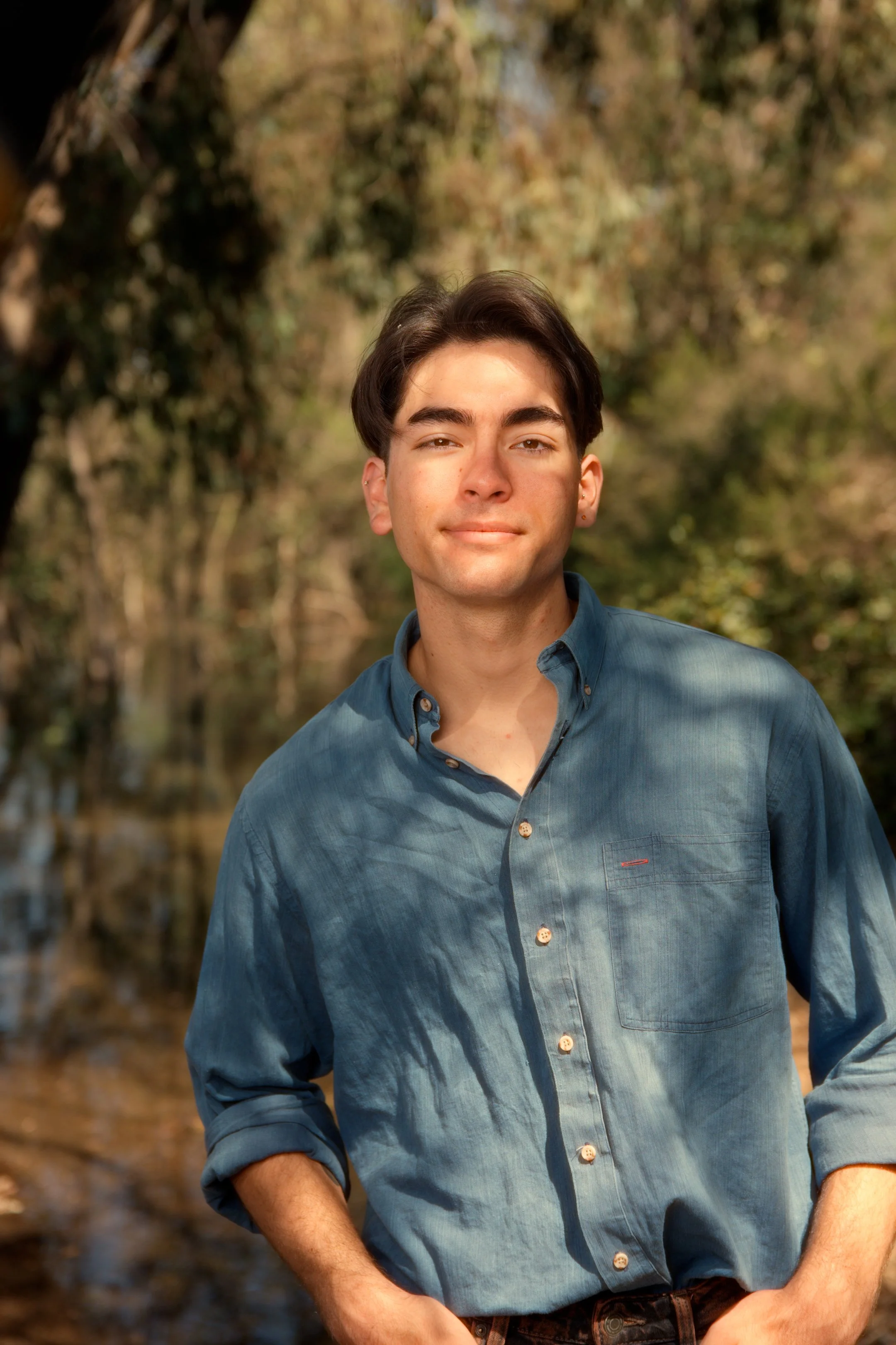 A young man with dark hair and fair skin standing outdoors in a wooded area with a creek in the background, wearing a blue button-up shirt with rolled sleeves.