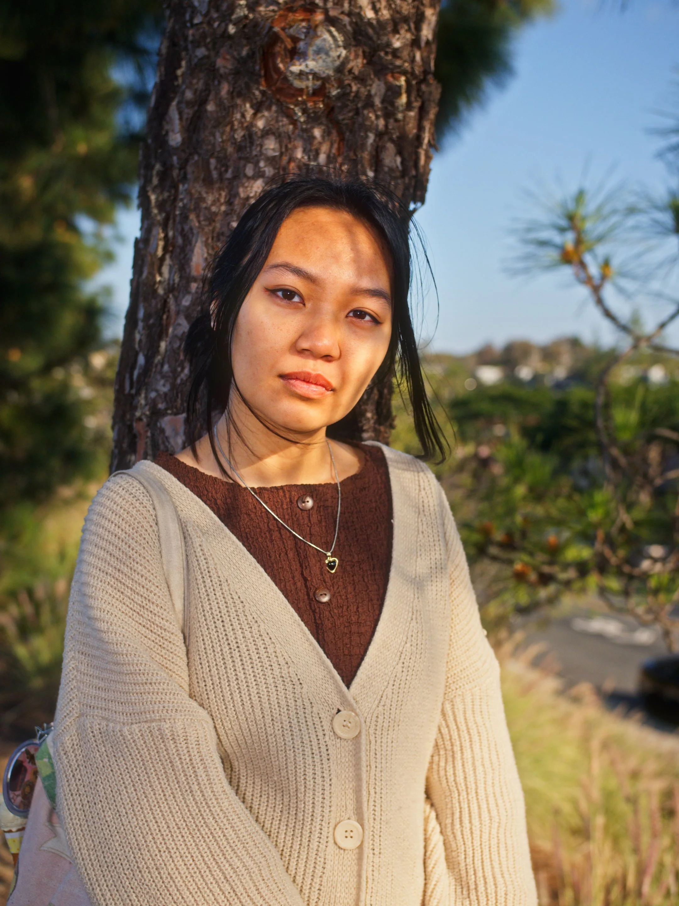 Young woman with black hair standing outdoors by a tree, wearing a beige cardigan over a brown top, with a necklace, looking at camera.