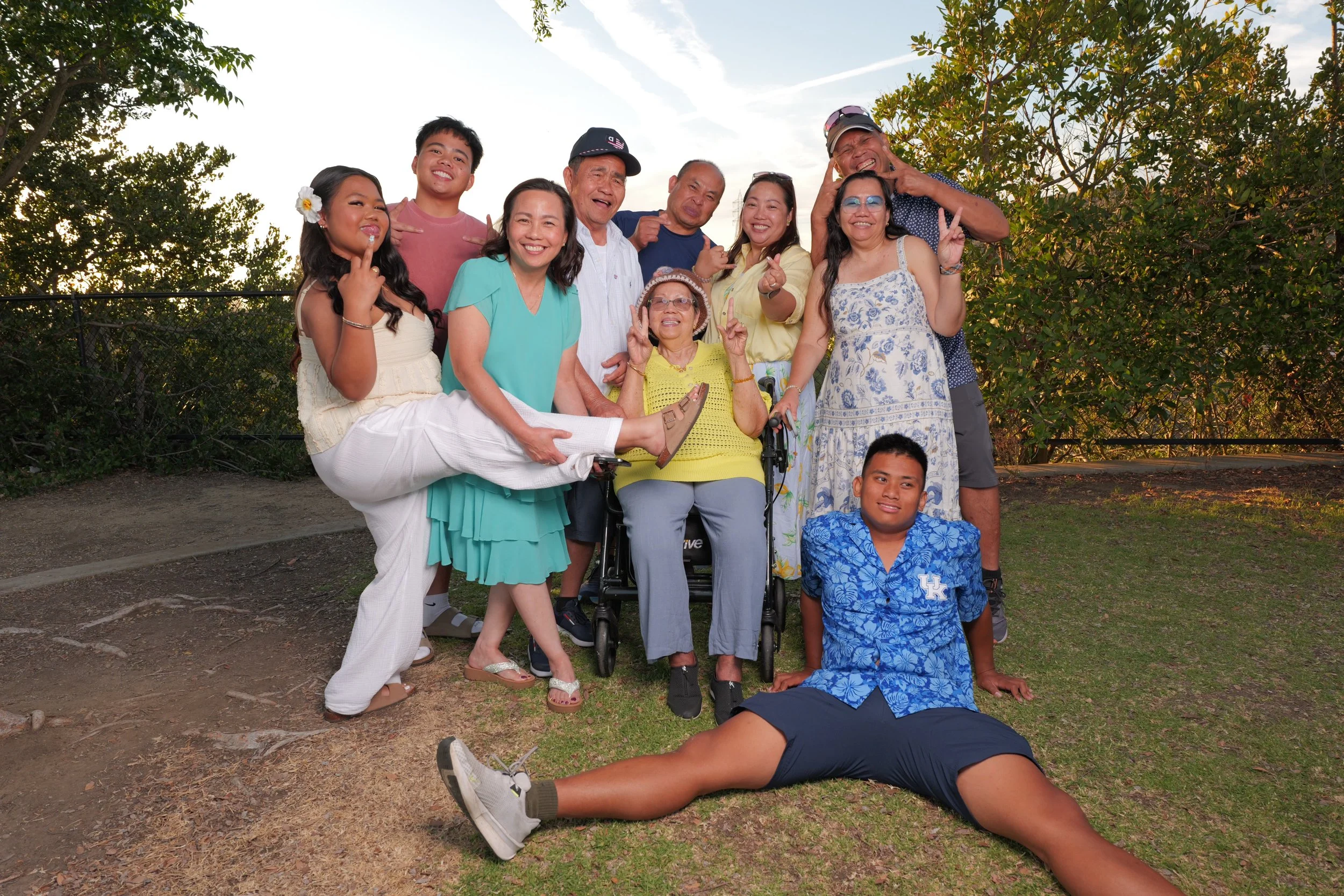 A group of ten diverse people posing outdoors, some making peace signs, smiling, and appearing cheerful during sunset in a park with trees in the background.