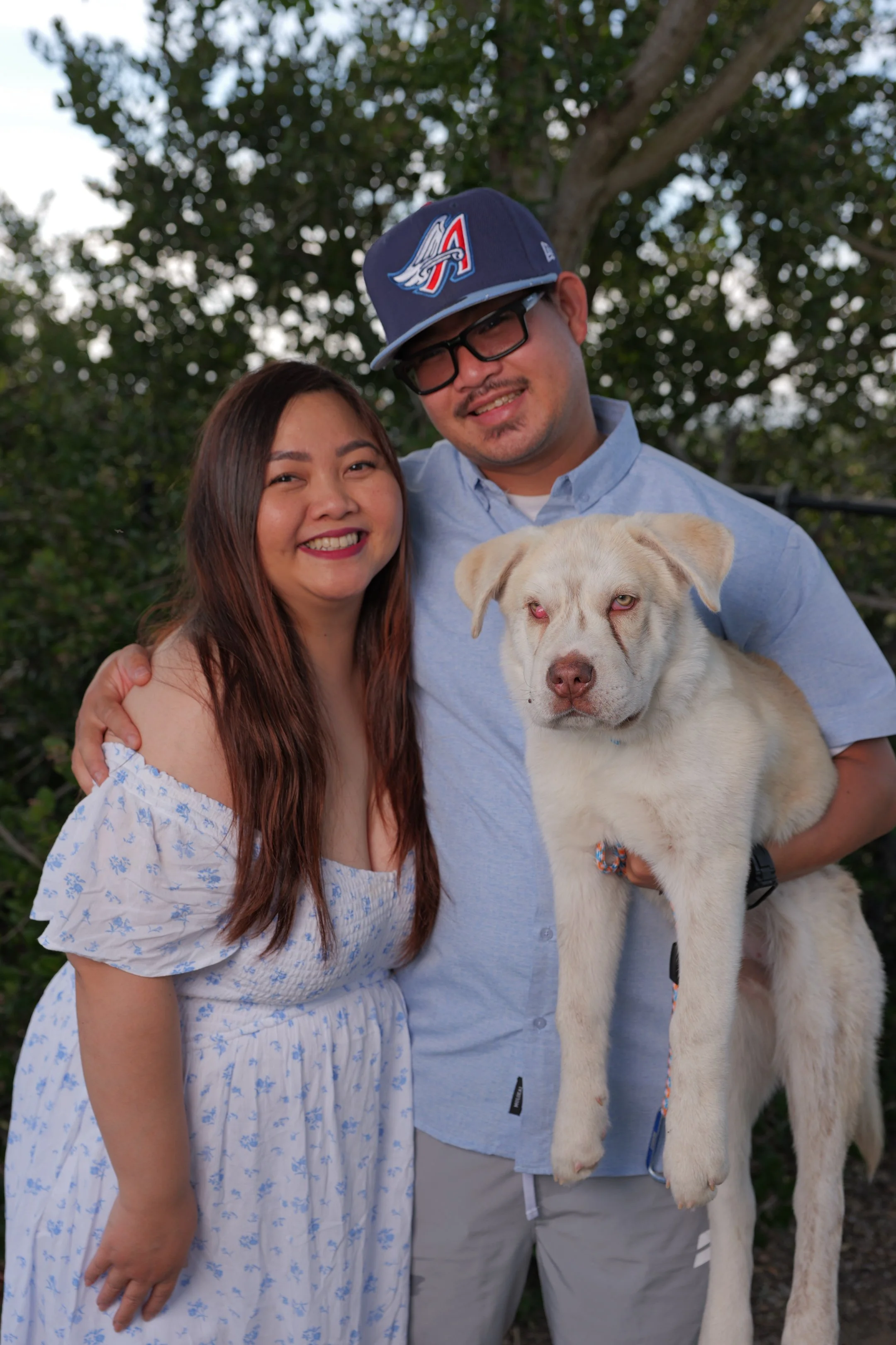 A smiling woman with long brown hair wearing a white off-shoulder dress standing next to a man in a blue shirt and cap holding a light-colored dog with red eyes in front of green trees.
