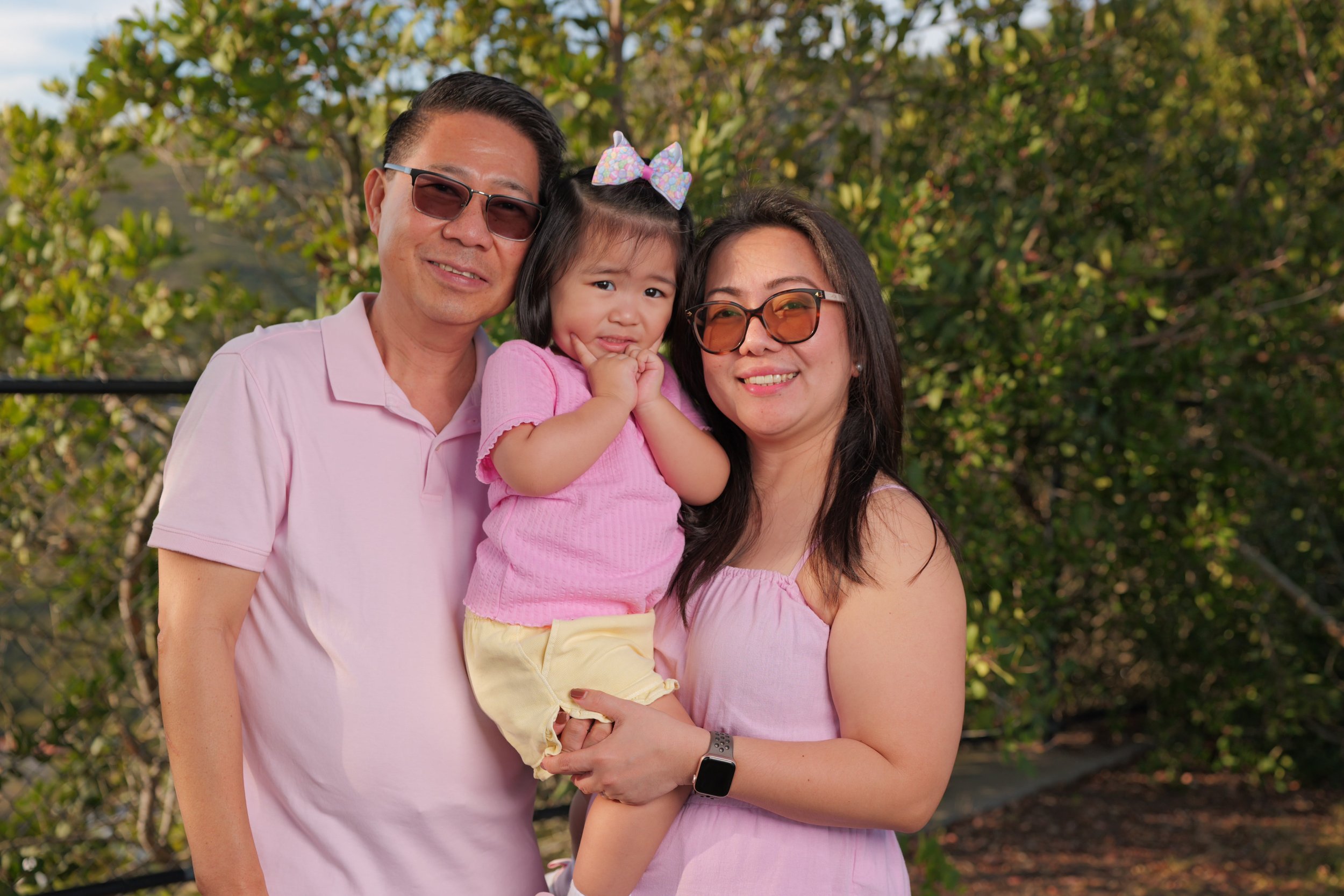 A smiling family of three, consisting of a man, woman, and young girl, outdoors with trees in the background. The man and woman are wearing glasses and pink tops, and the girl is wearing a pink shirt with a rainbow-colored bow in her hair. The woman 