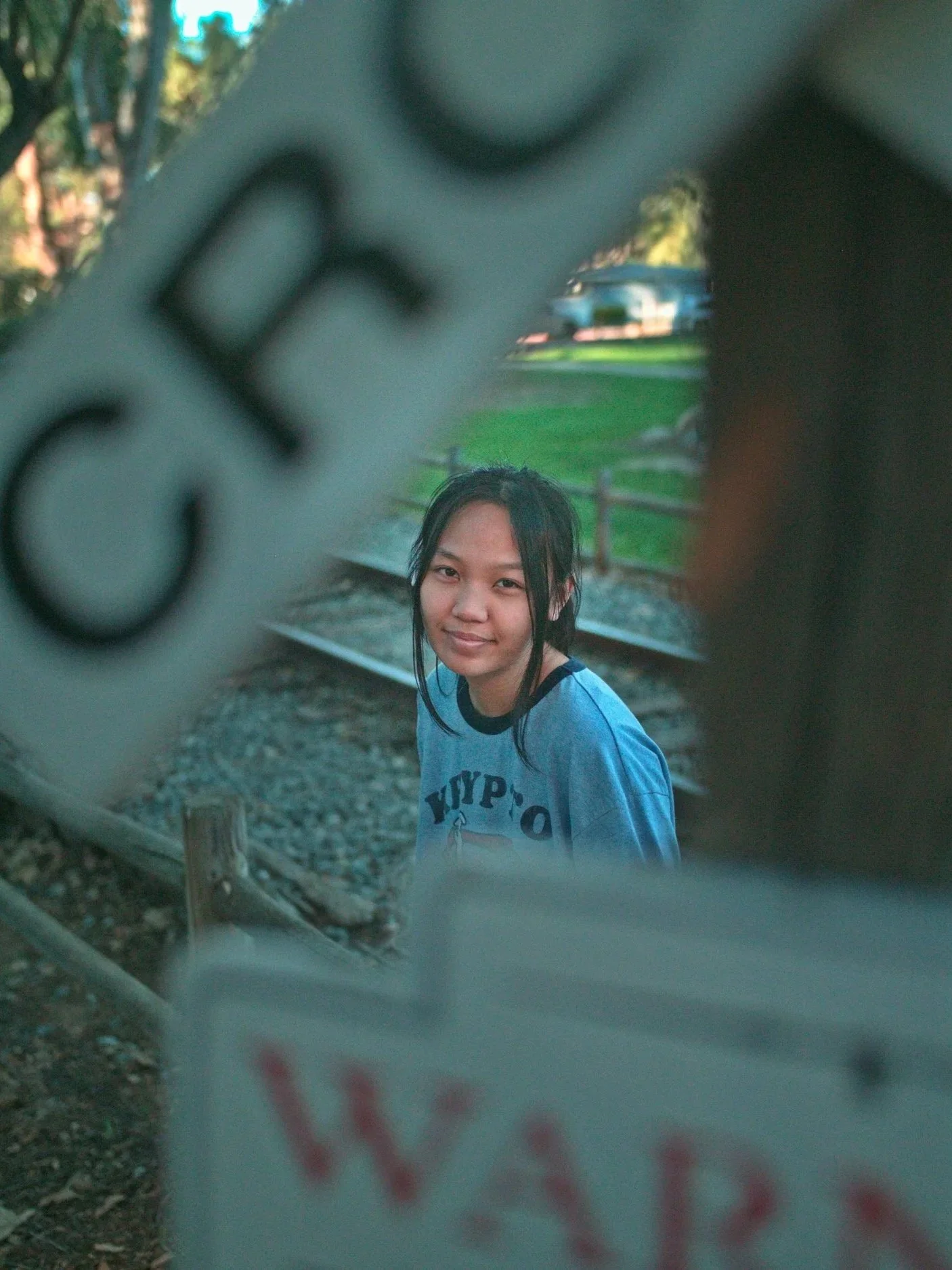 A young woman with black hair and a blue shirt with black trim, standing outdoors near train tracks, smiling and looking at the camera through a hole in a fence or barrier.