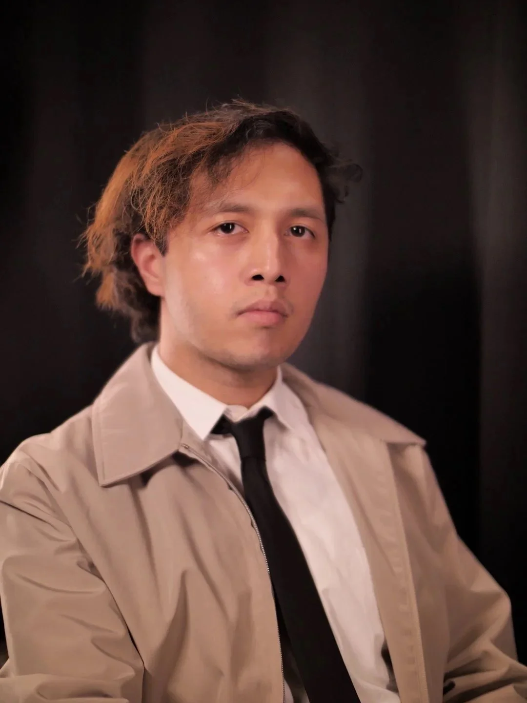 A young man with medium-length, tousled hair and light skin, wearing a beige trench coat over a white shirt and black tie, standing against a dark background.