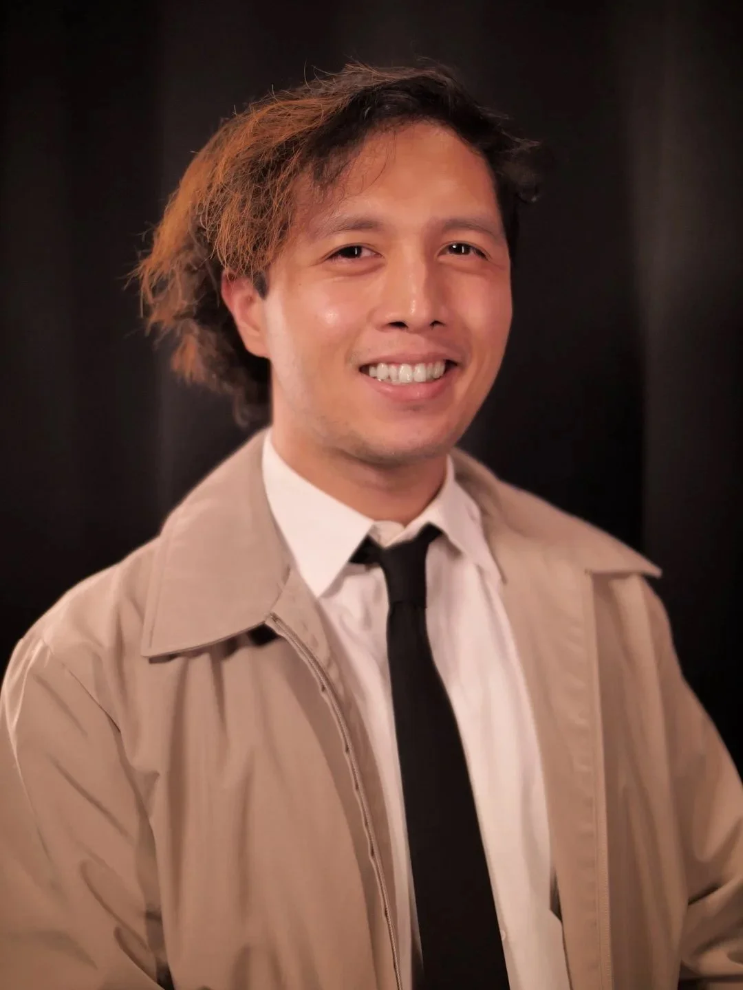 A smiling man with slightly wavy hair wearing a tan jacket, white shirt, and black tie standing against a dark background.