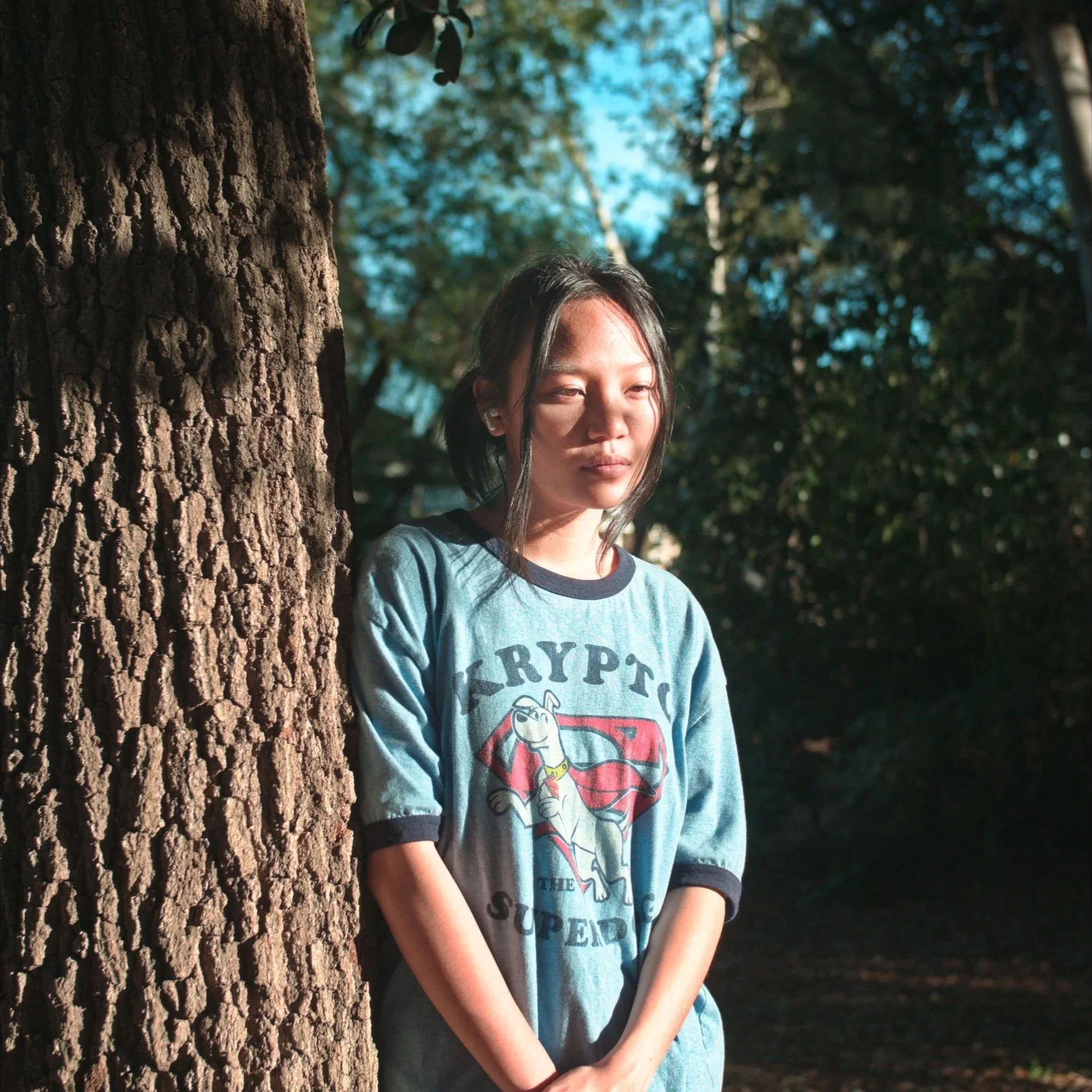 A young woman standing outdoors in front of a tree, with sunlight illuminating her face, wearing a blue T-shirt with Superman and Skepti logo, with a forest background.