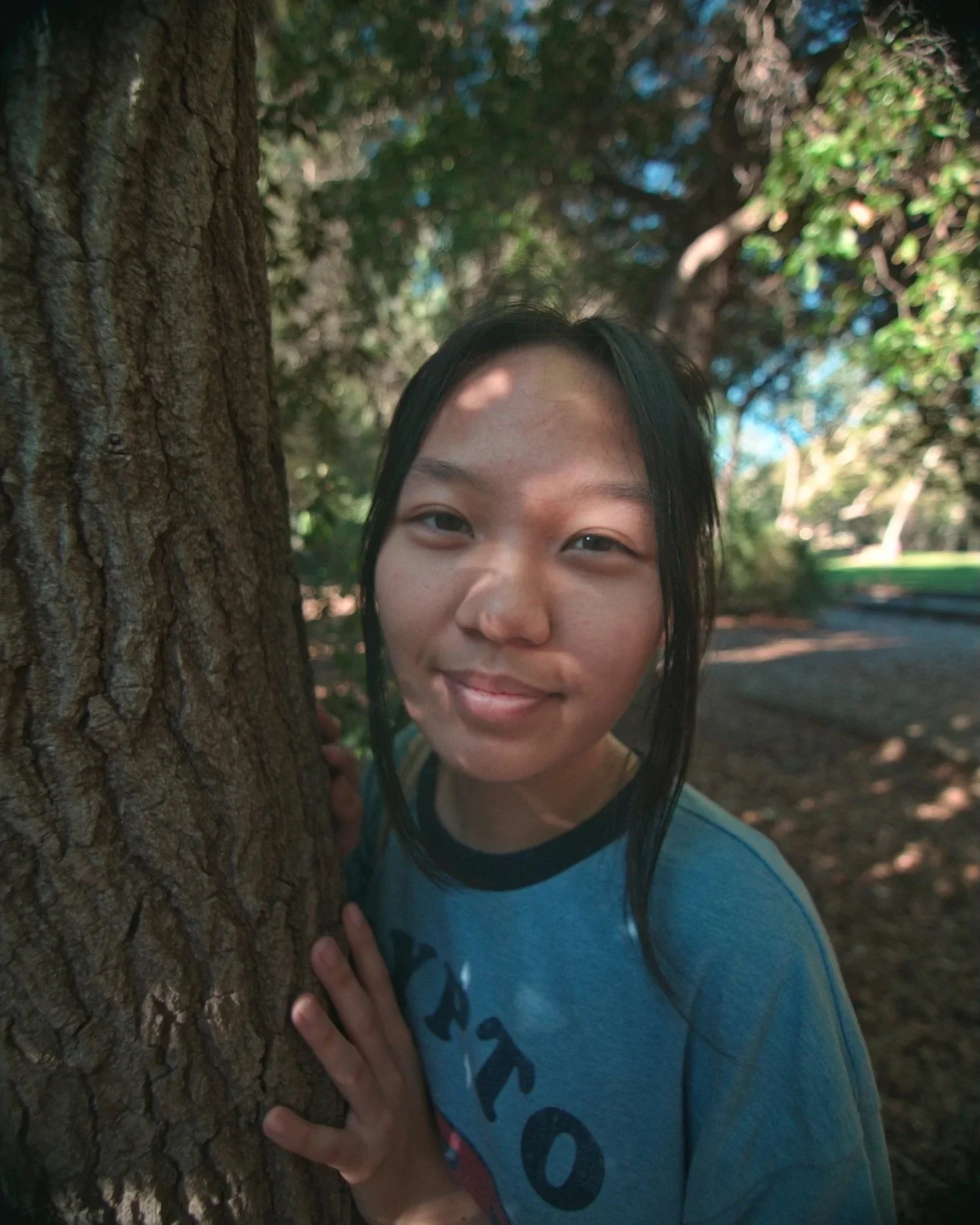 A girl with black hair peeking from behind a tree with a smile in a park.