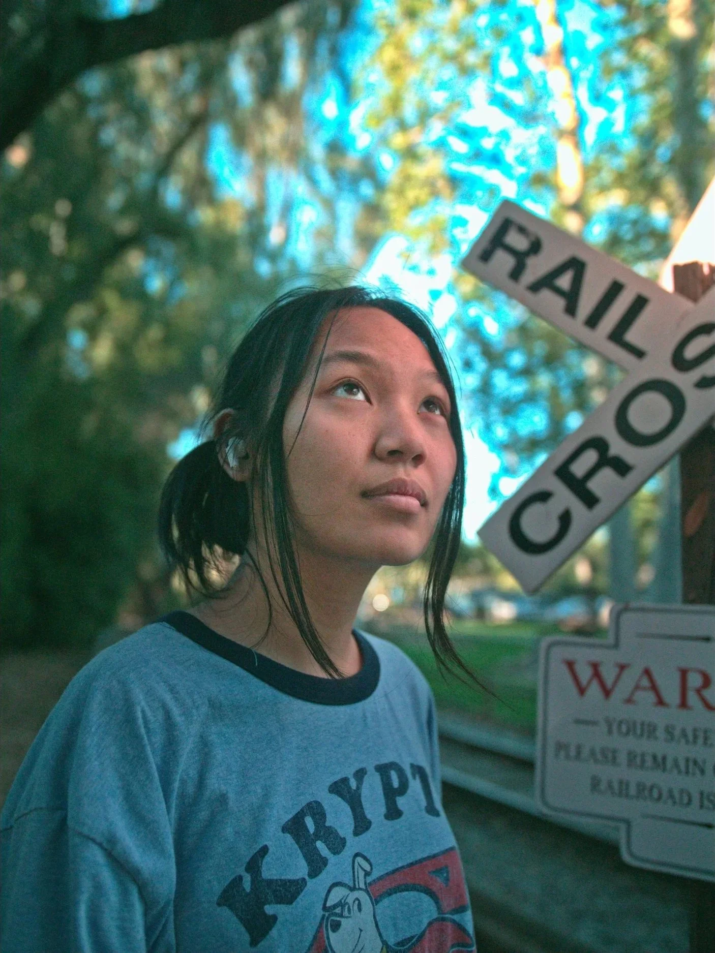 A young woman with black hair in a ponytail looking upward outdoors, standing near a railroad crossing sign, with trees and blue sky in the background.