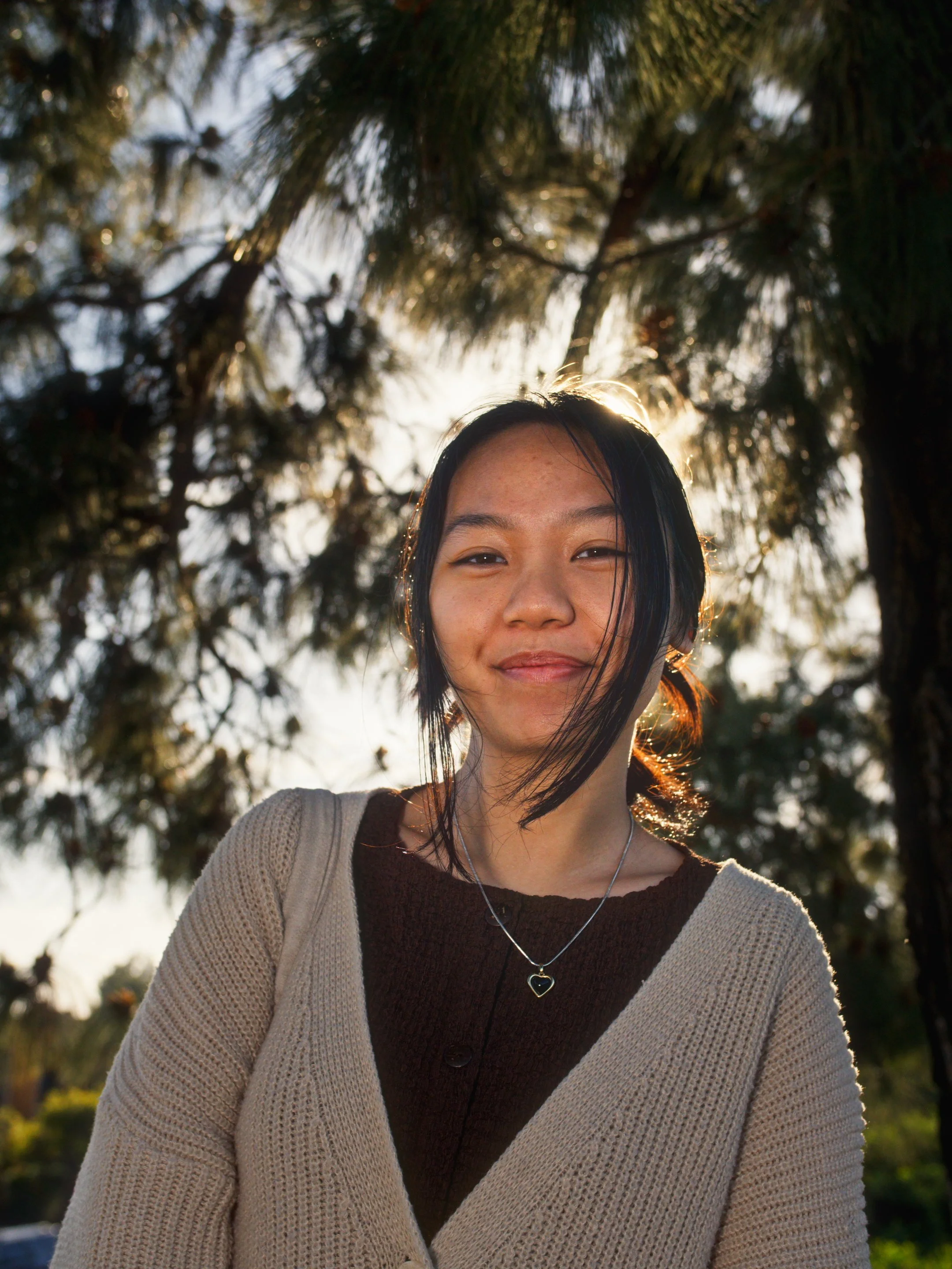 A young woman standing outdoors with trees in the background, backlit by the sun, smiling softly.