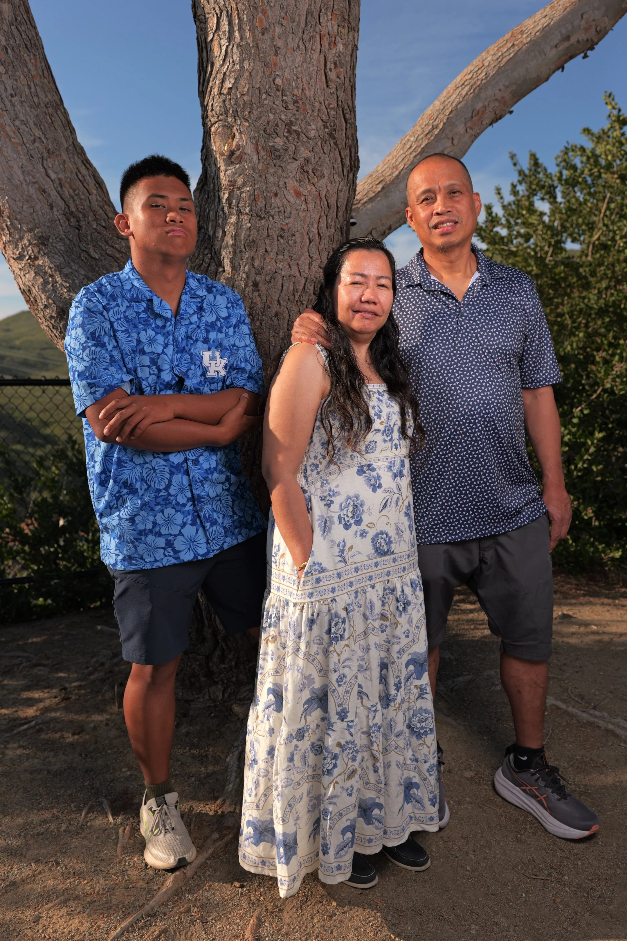 A family of three stands outdoors next to a large tree, with a clear blue sky in the background. The son on the left is wearing a blue Hawaiian shirt with a University of Kentucky logo, black shorts, and sneakers. The mother in the middle is wearing 