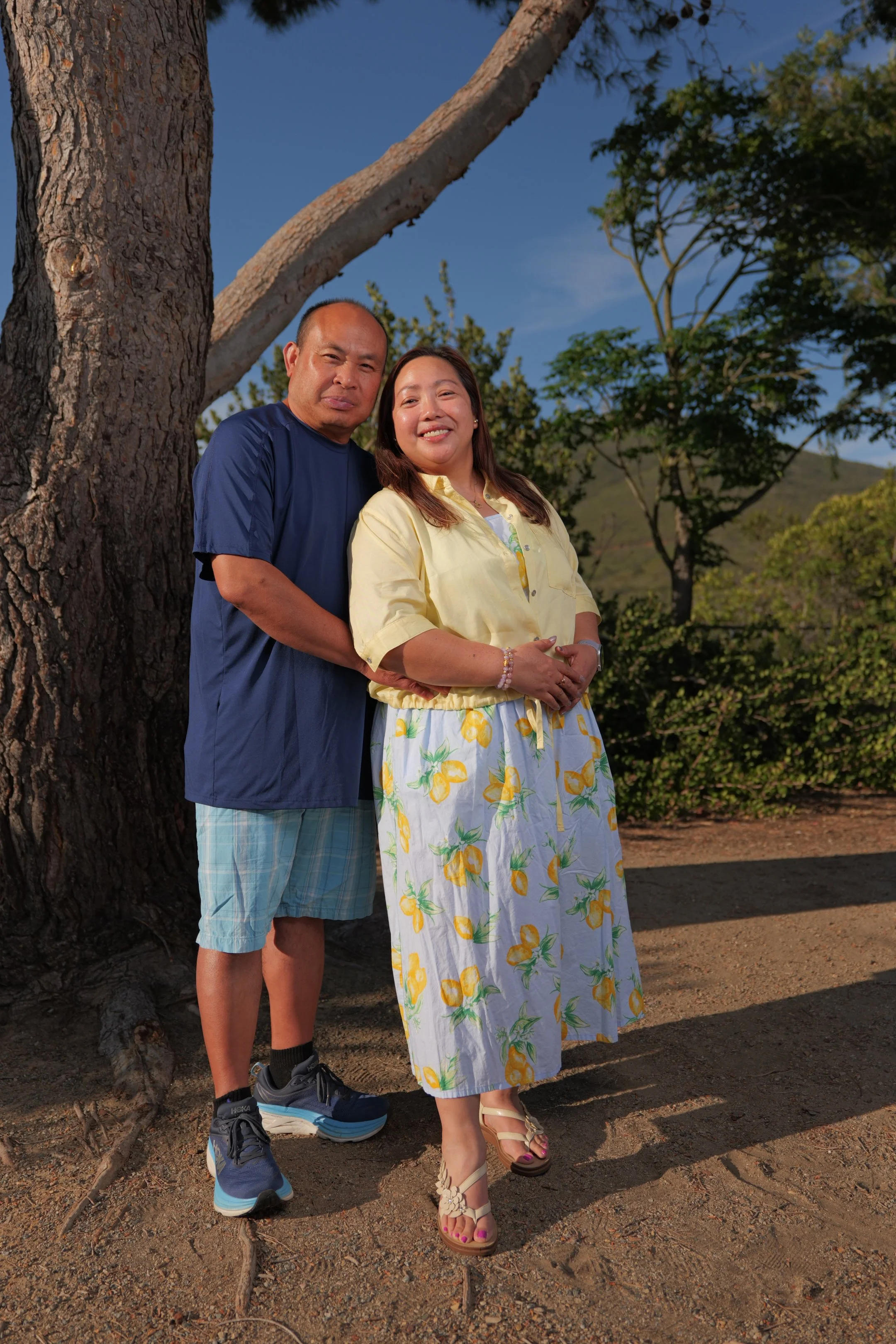 A man and woman standing outdoors next to a large tree, smiling at the camera, with trees and a hill in the background on a sunny day.