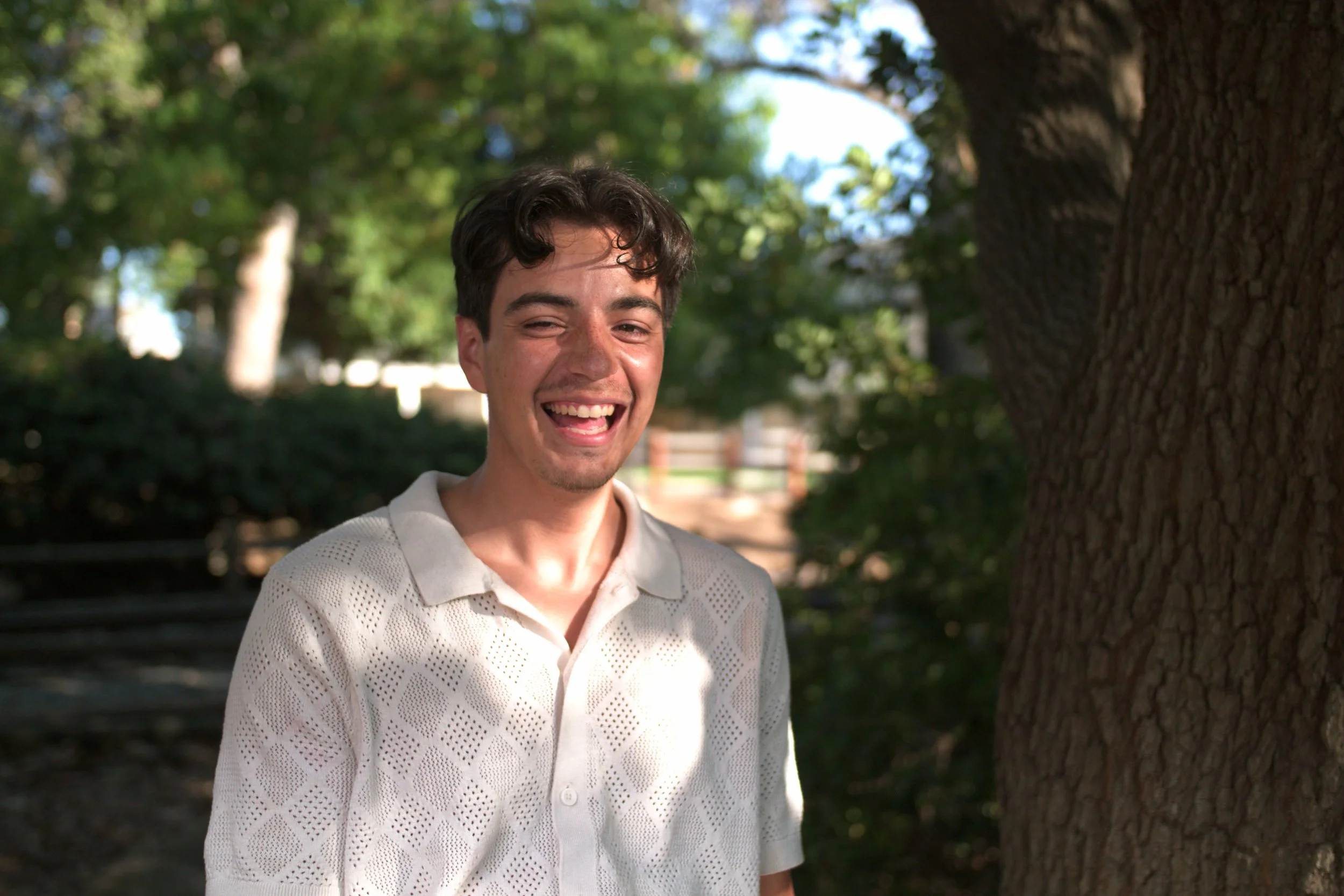 A young man smiling outdoors next to a large tree with sunlight and green foliage in the background.