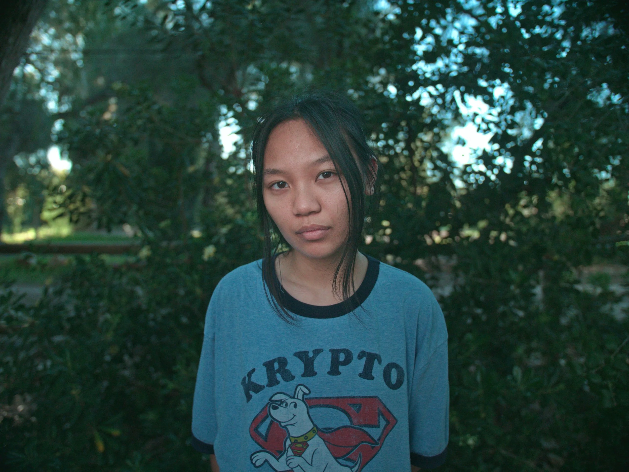 A young woman with straight black hair wearing a gray T-shirt with a cartoon dog and Superman logo, standing outdoors in a green, leafy park.