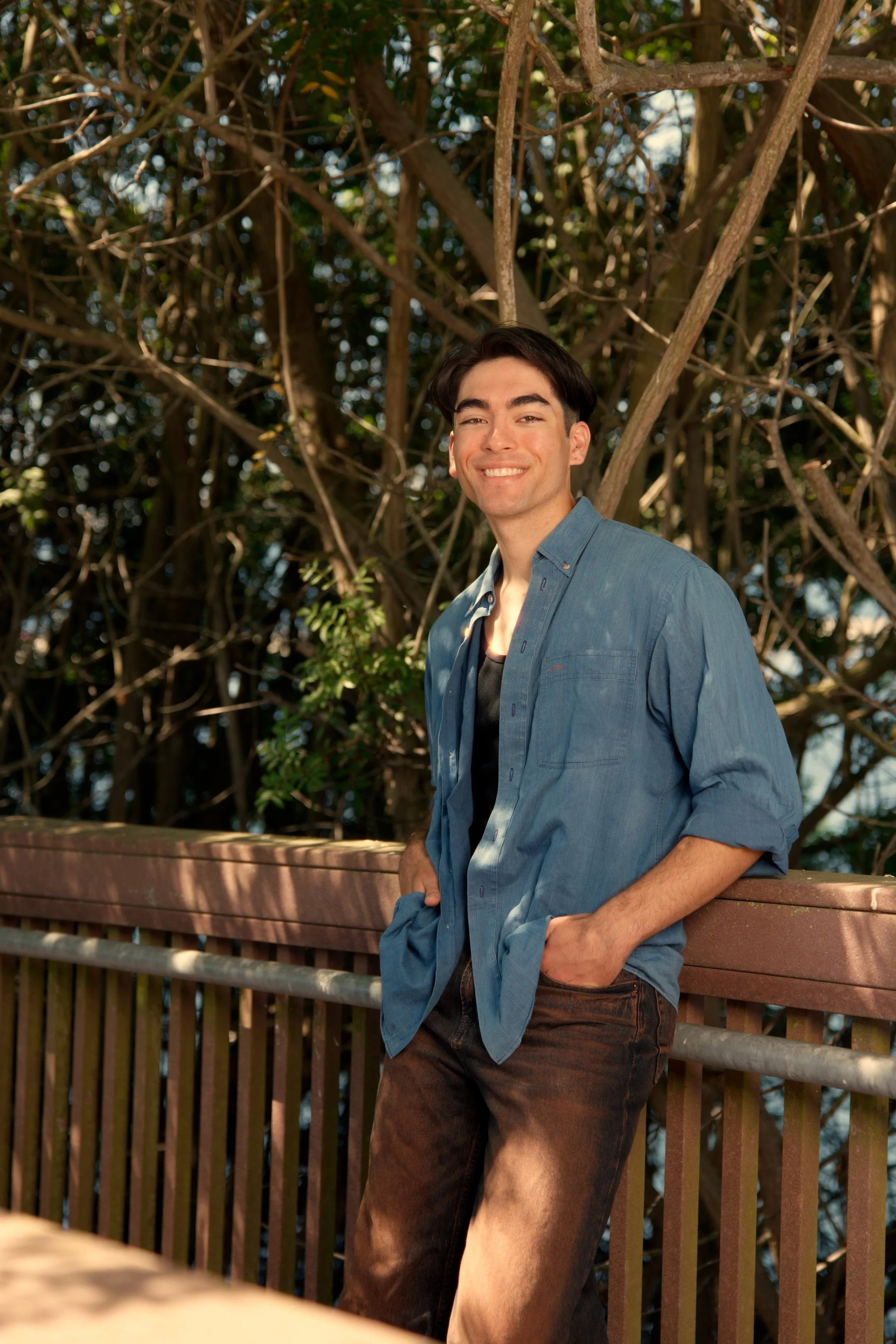 A young man with dark hair and a smile, wearing a blue shirt and dark jeans, leaning casually against a wooden railing on an outdoor balcony with tree branches in the background.