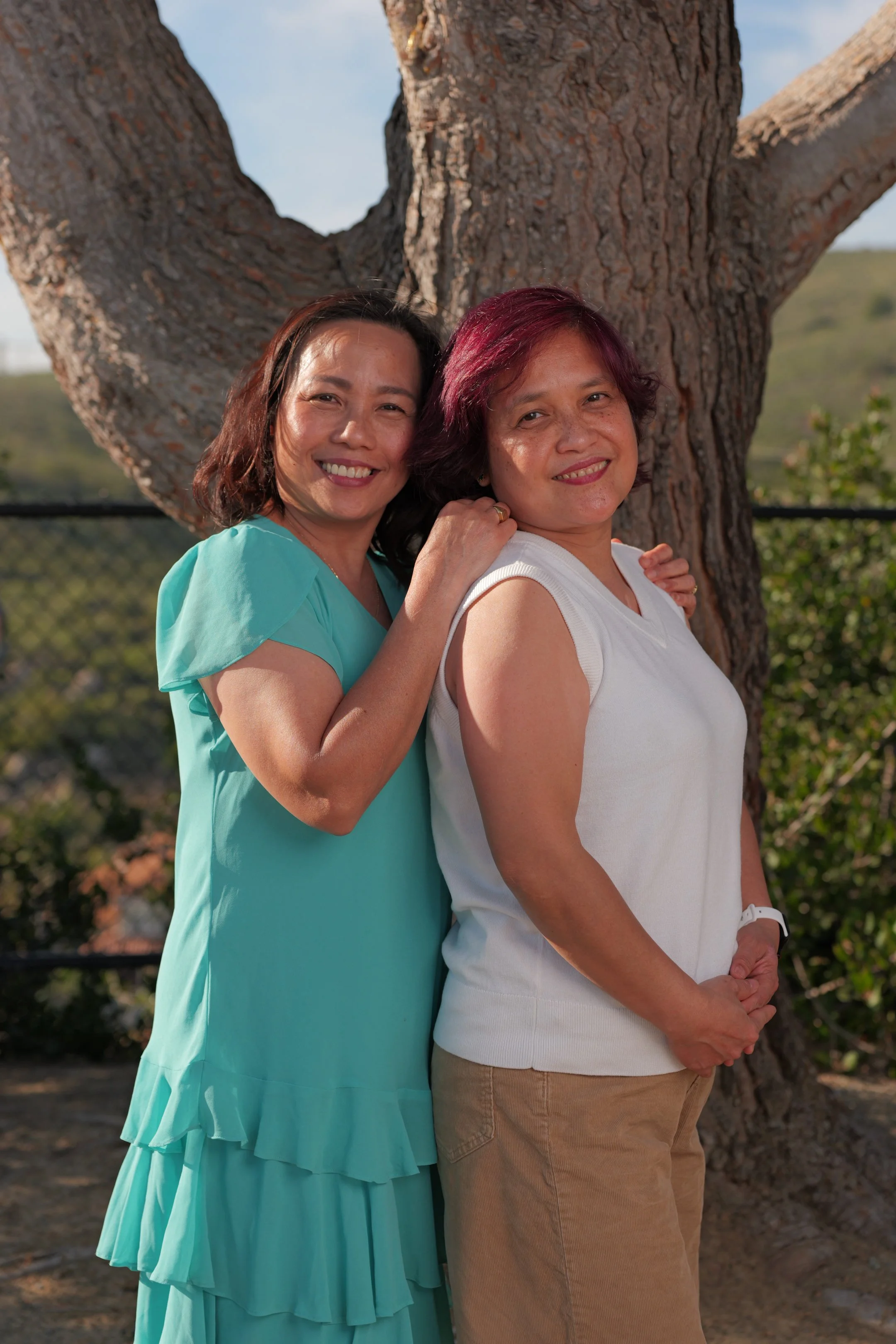 Two women standing outdoors near a large tree, smiling at the camera, with a scenic background of hills and sky.