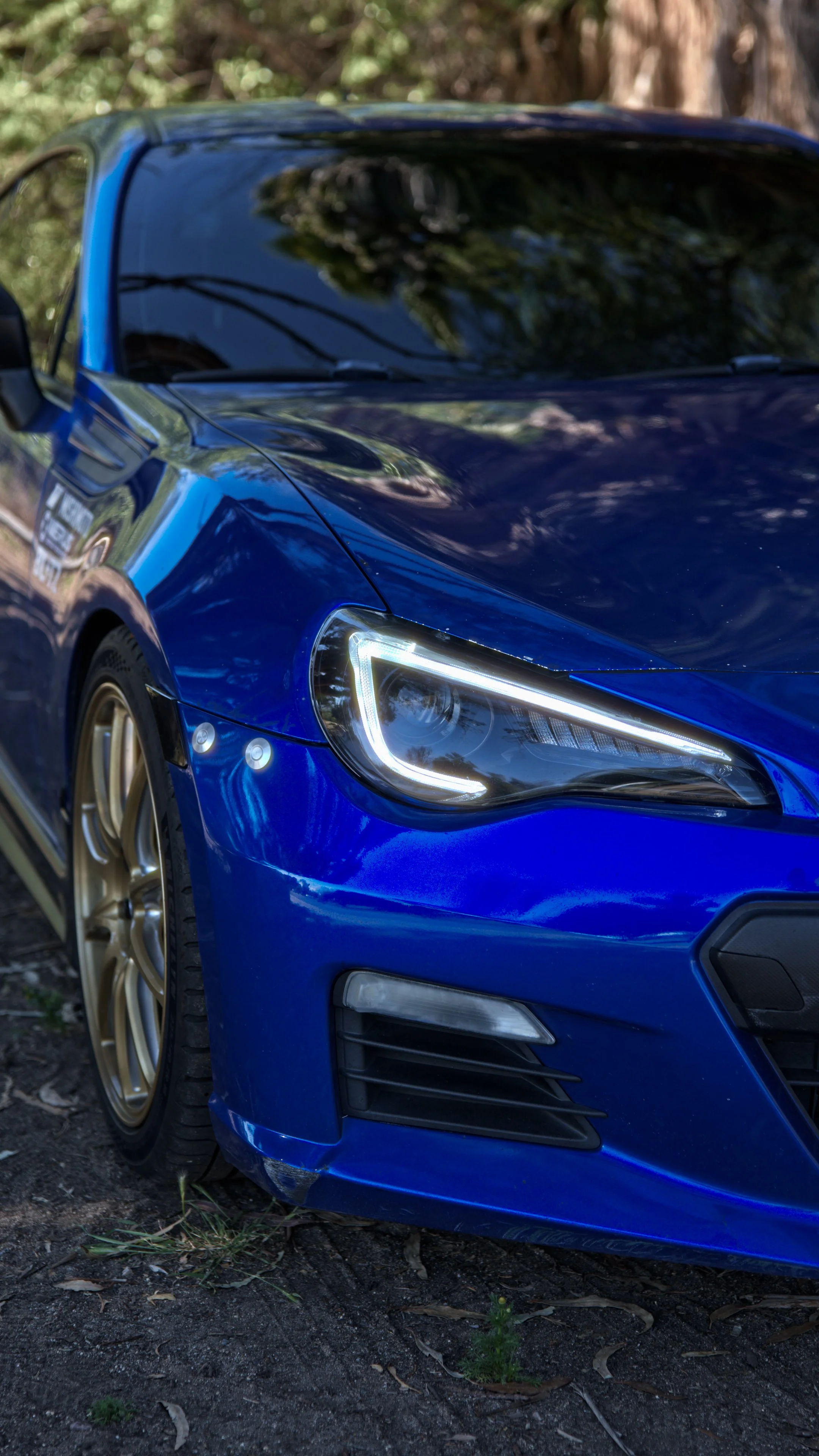 Close-up of a blue sports car with headlights on, parked outdoors on dirt with trees reflected in the windshield.
