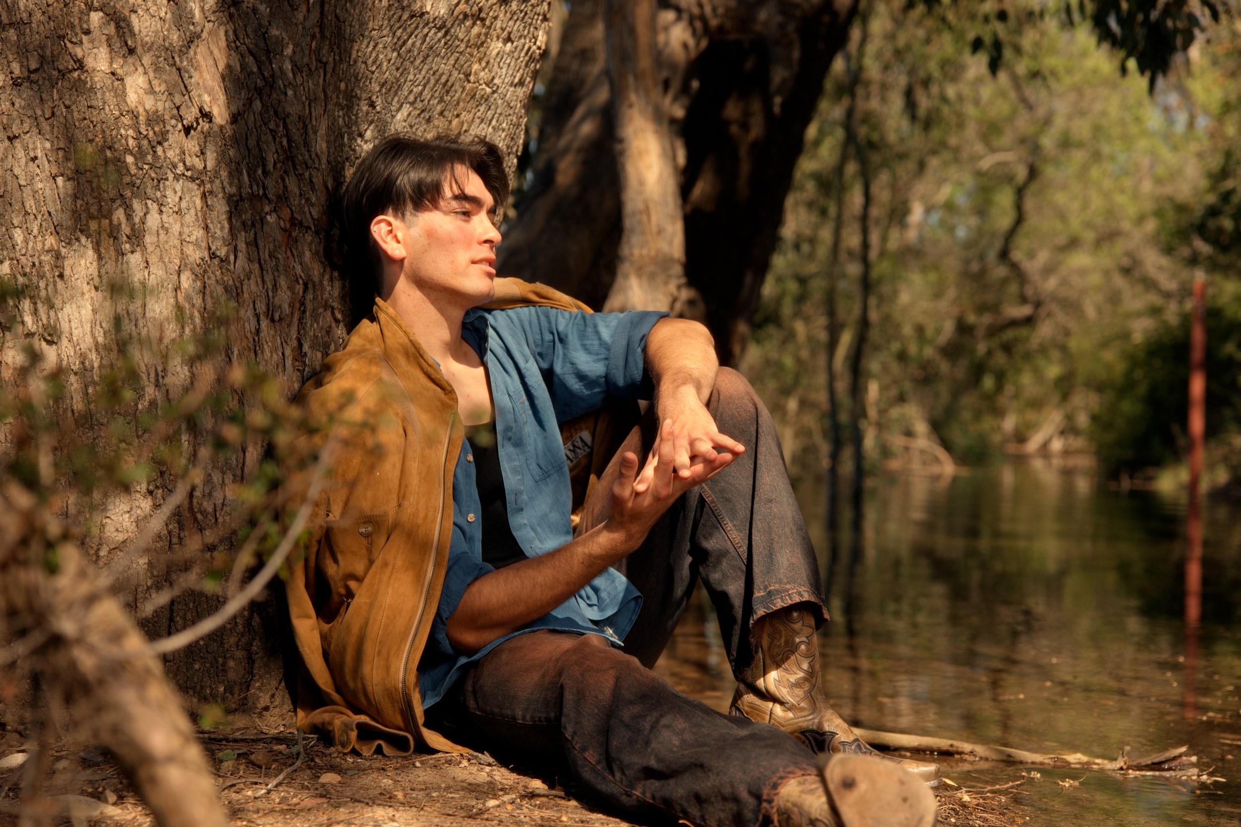 A young man with dark hair, wearing a brown jacket, blue shirt, and jeans, sitting on the ground and leaning against a large tree by a river, appearing contemplative.