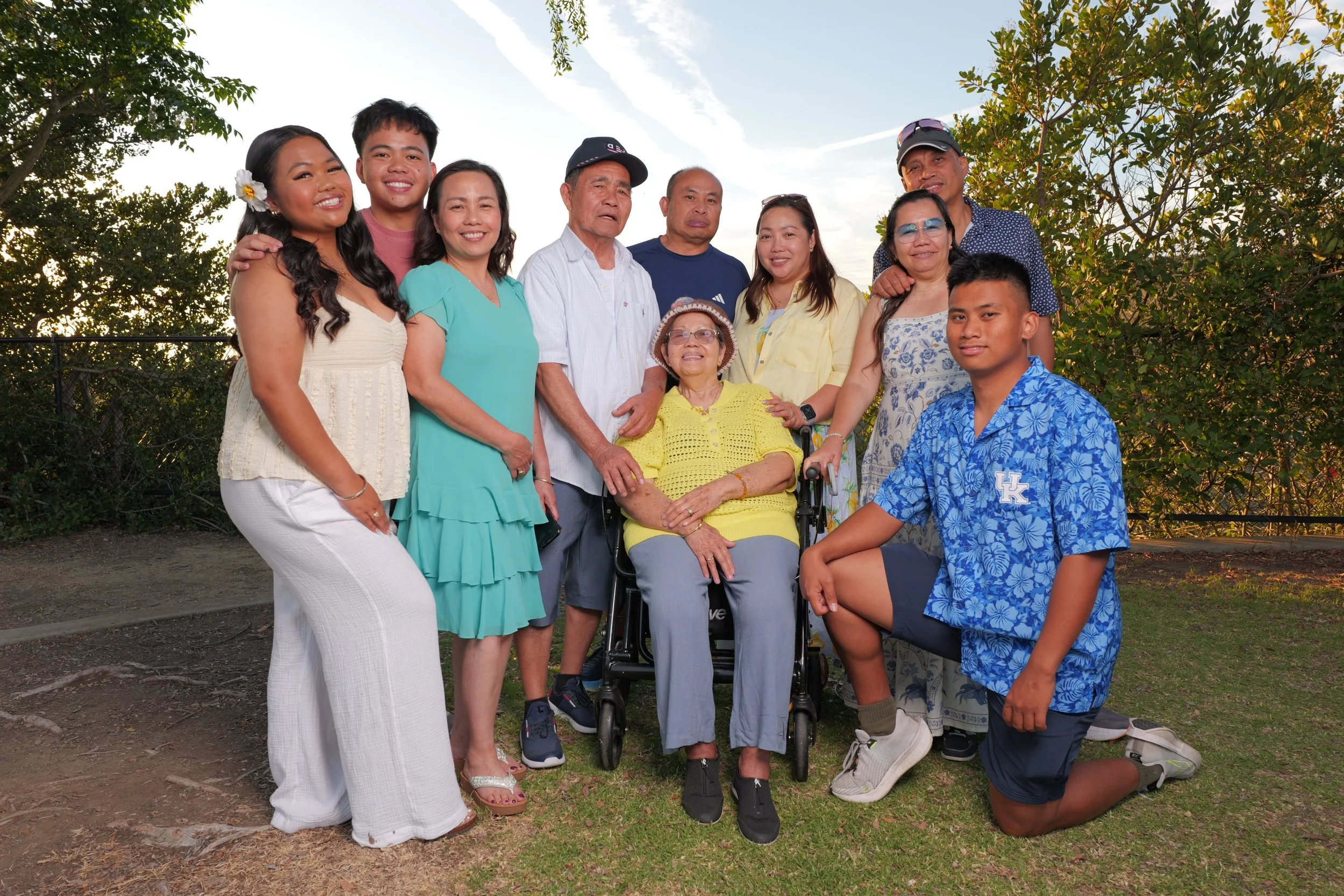 Family group photo outdoors with trees and sky in the background, featuring multiple generations with an elderly woman in a wheelchair at the center.