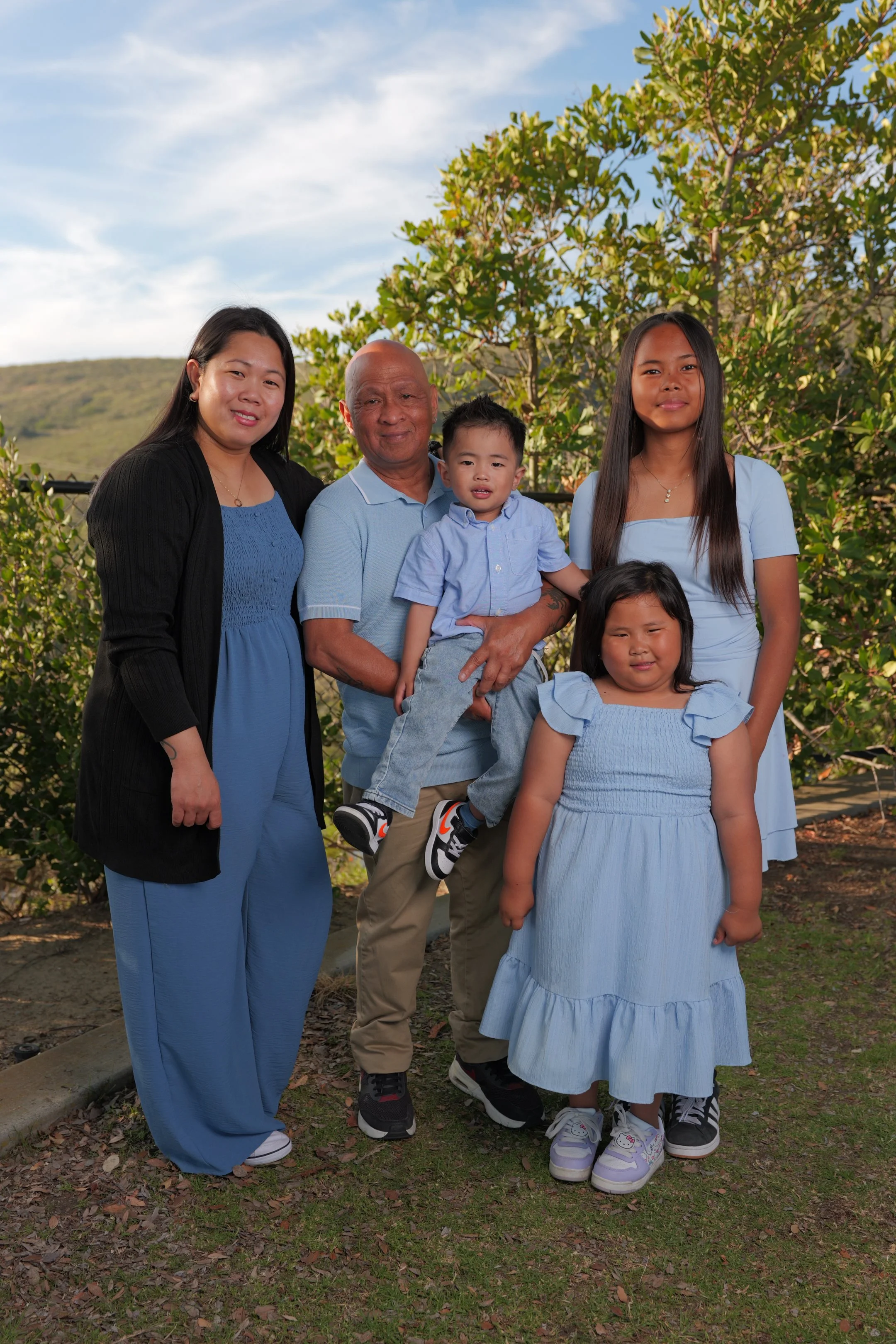 A family of five standing outdoors in front of greenery, with a mountain and blue sky in the background, dressed in blue clothing.