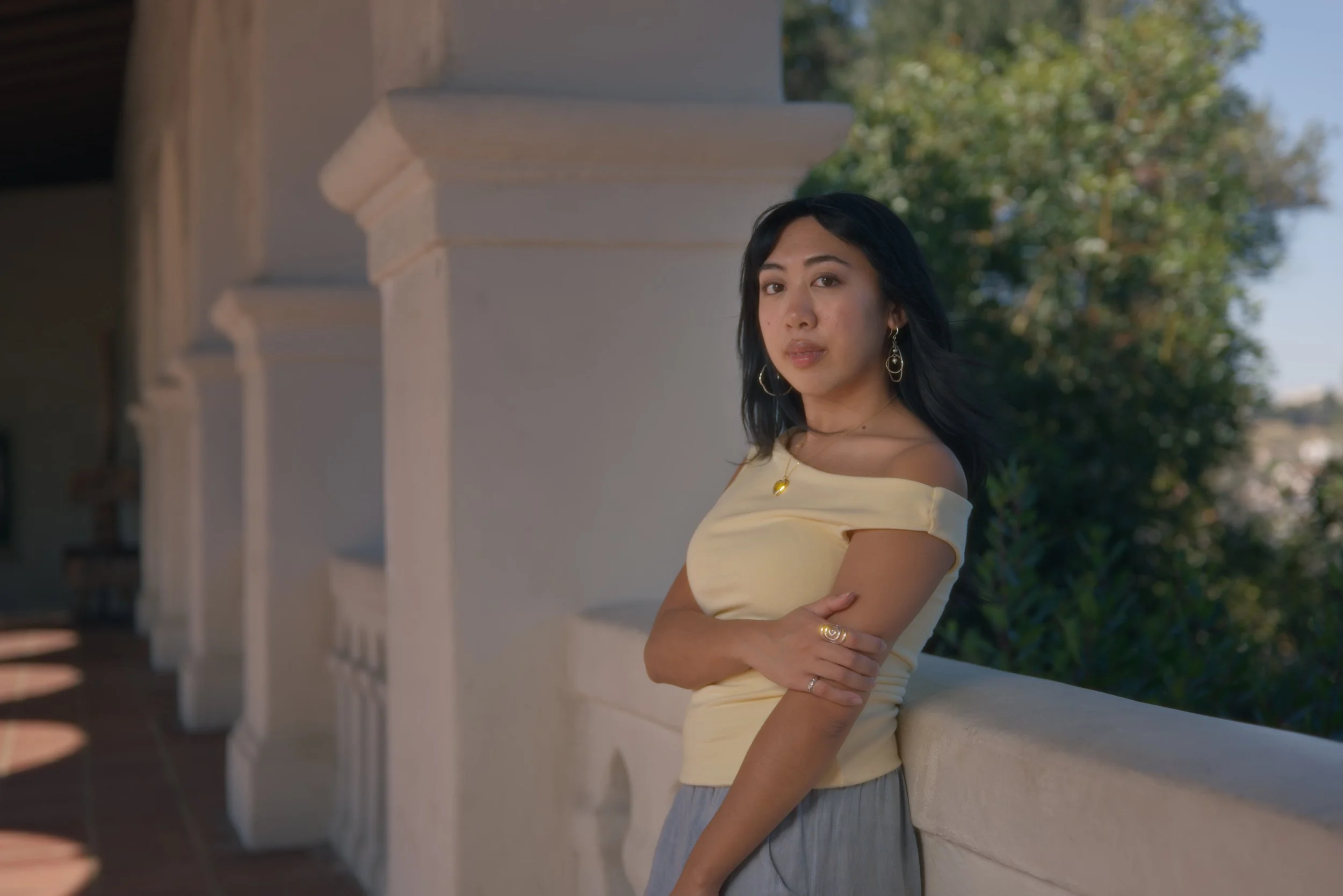 A woman with black hair wearing a yellow off-shoulder top and gray skirt, standing on a balcony with white architectural details and greenery in the background.