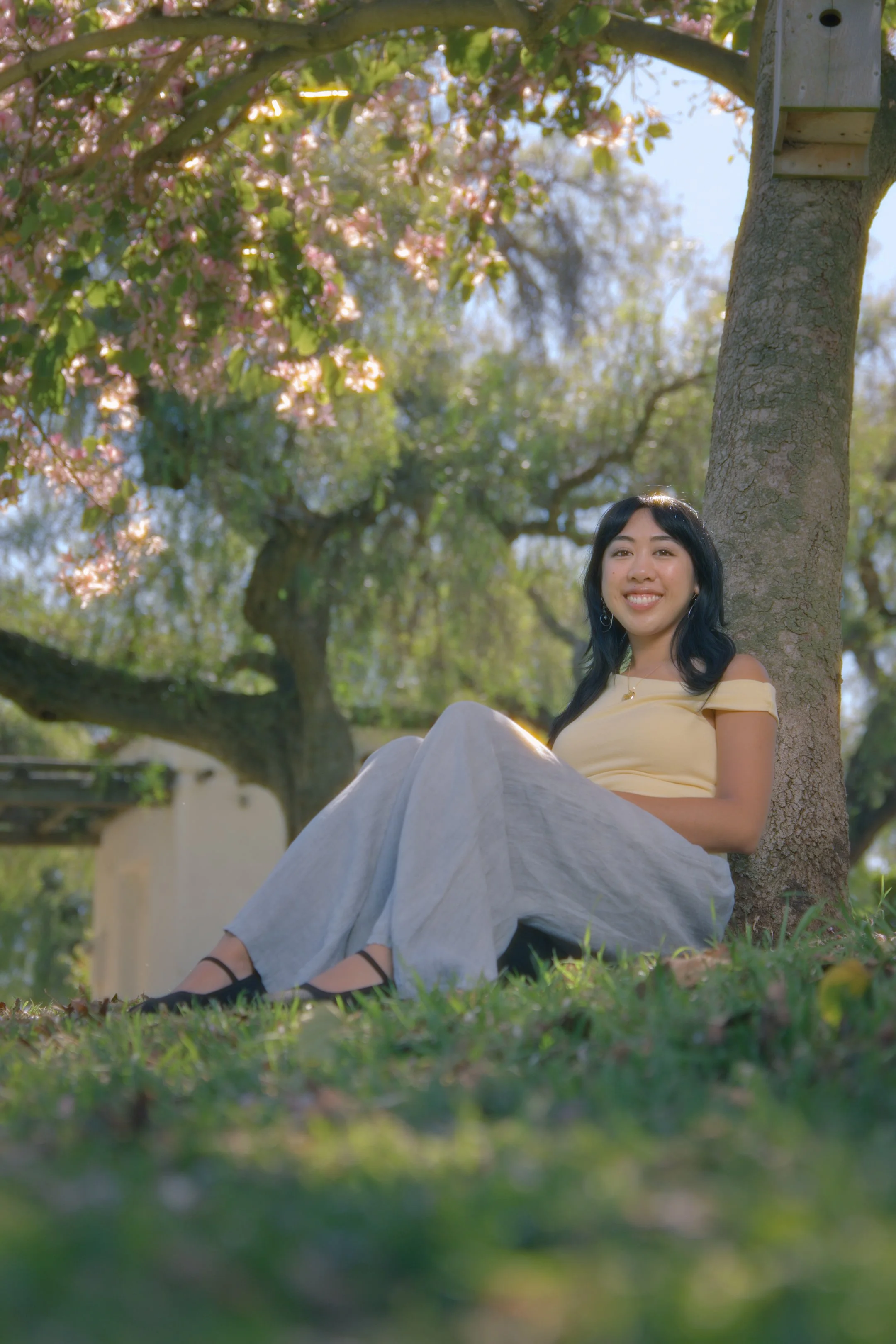 A young woman sitting under a tree in a park, smiling at the camera, with blooming flowers and lush green foliage around her.