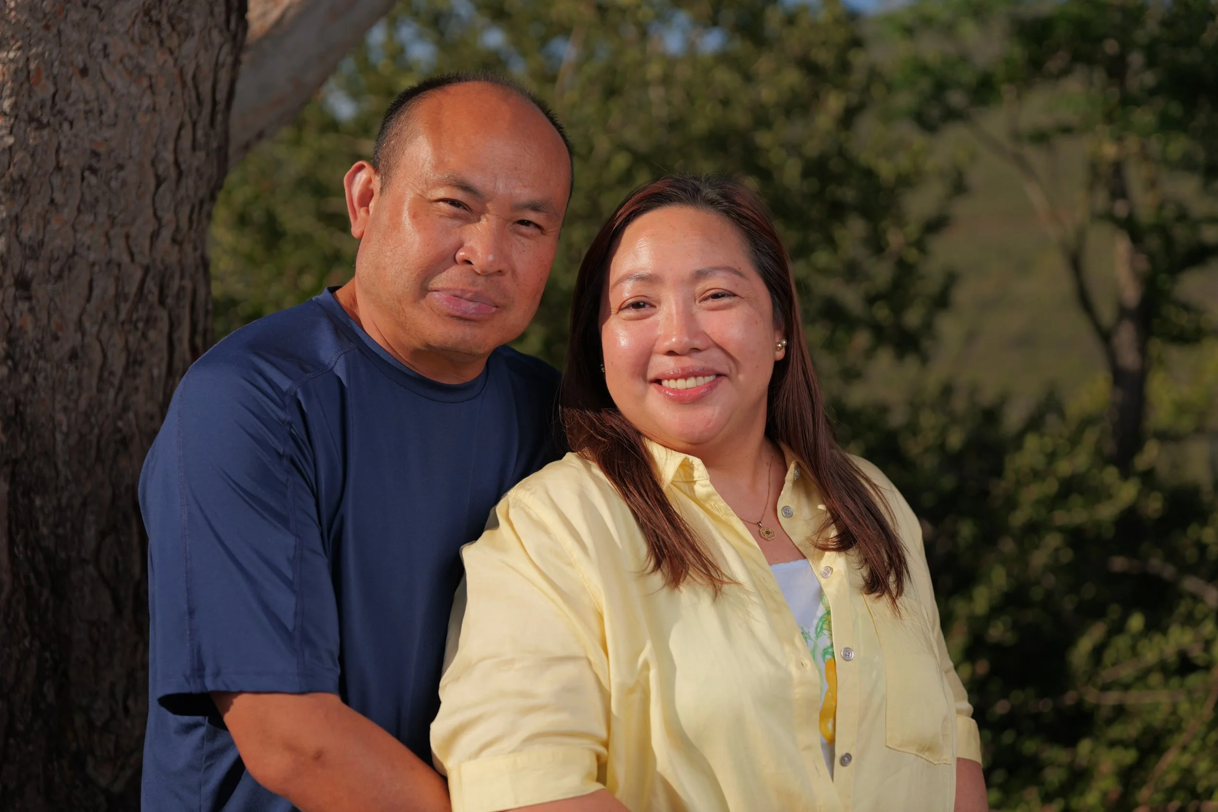 A man and woman smiling outdoors beside a tree with a green forest background.