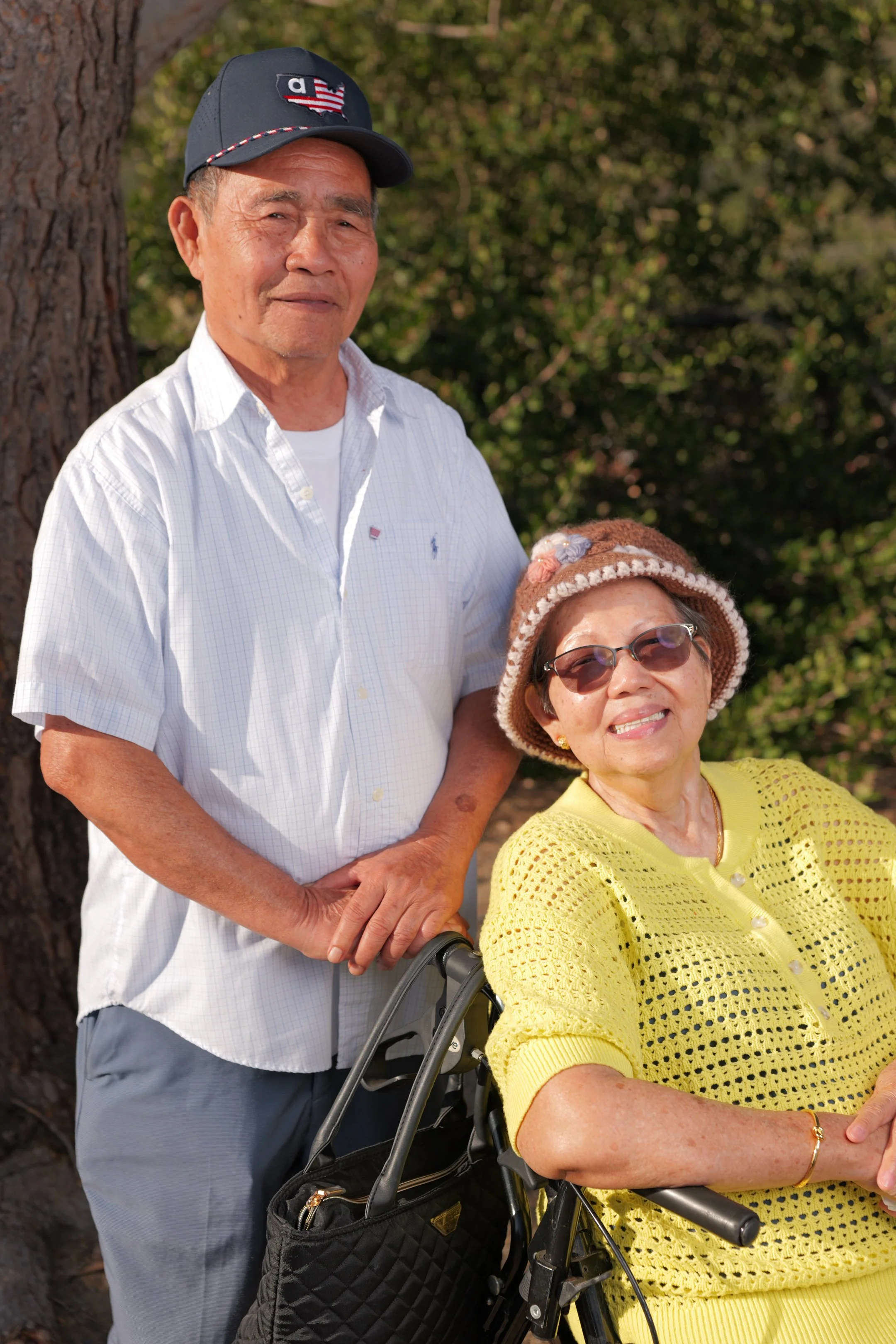 An elderly man with a baseball cap standing beside an elderly woman in a wheelchair, wearing sunglasses and a yellow outfit, outdoors in front of trees.