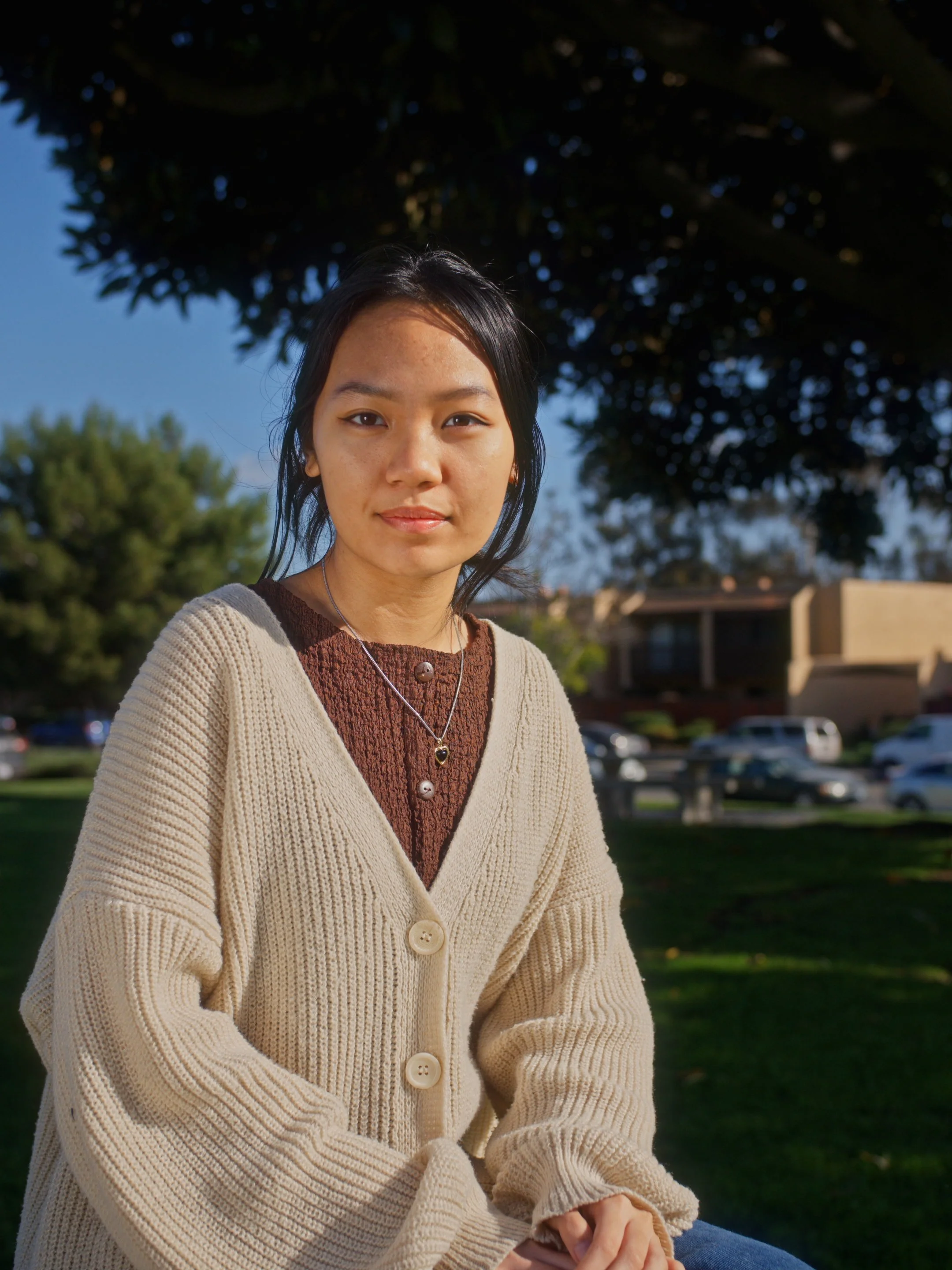 A young woman sitting outdoors on a sunny day with a tree and a residential area in the background.