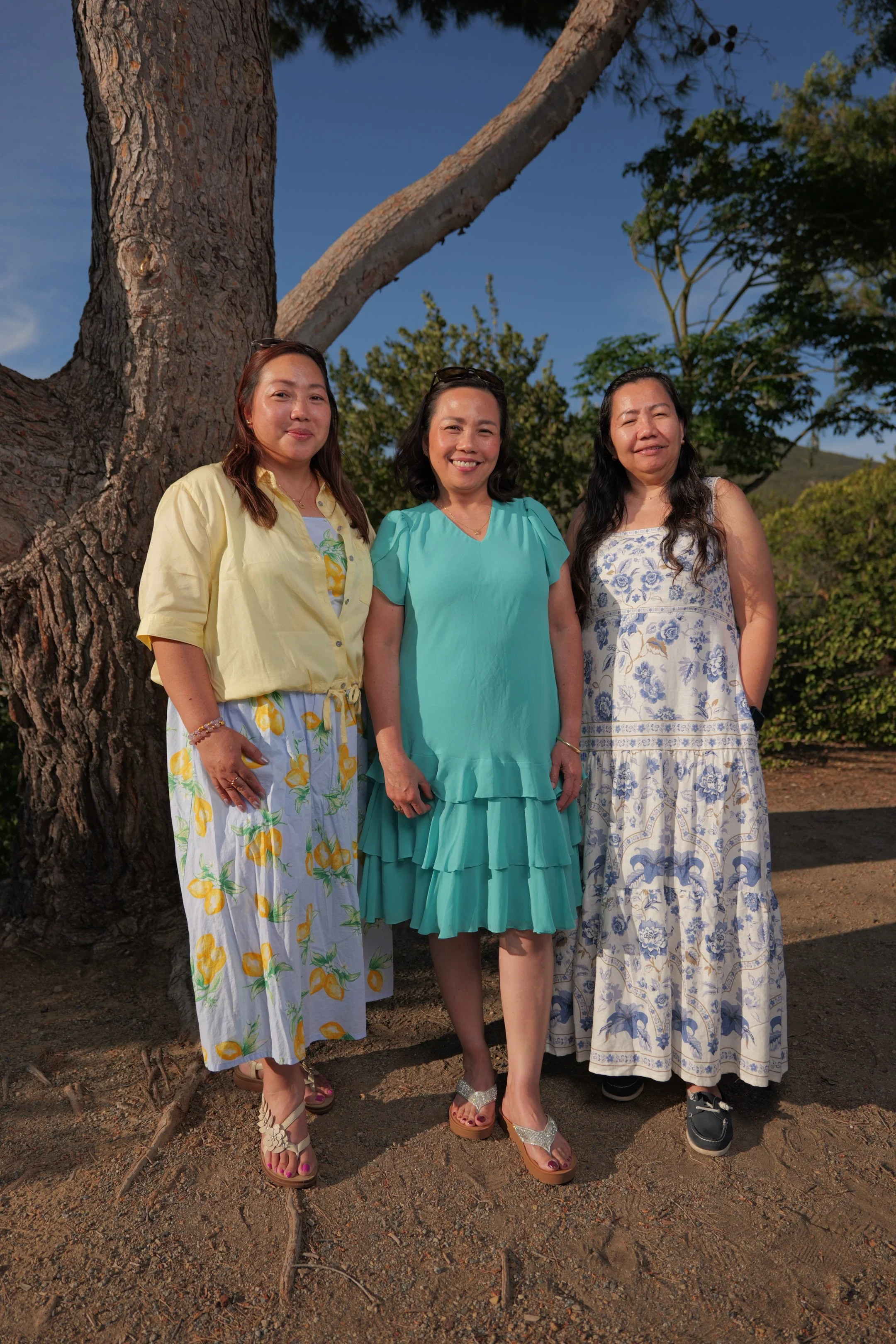 Three women standing outdoors in front of a large tree and green foliage, smiling at the camera on a sunny day.