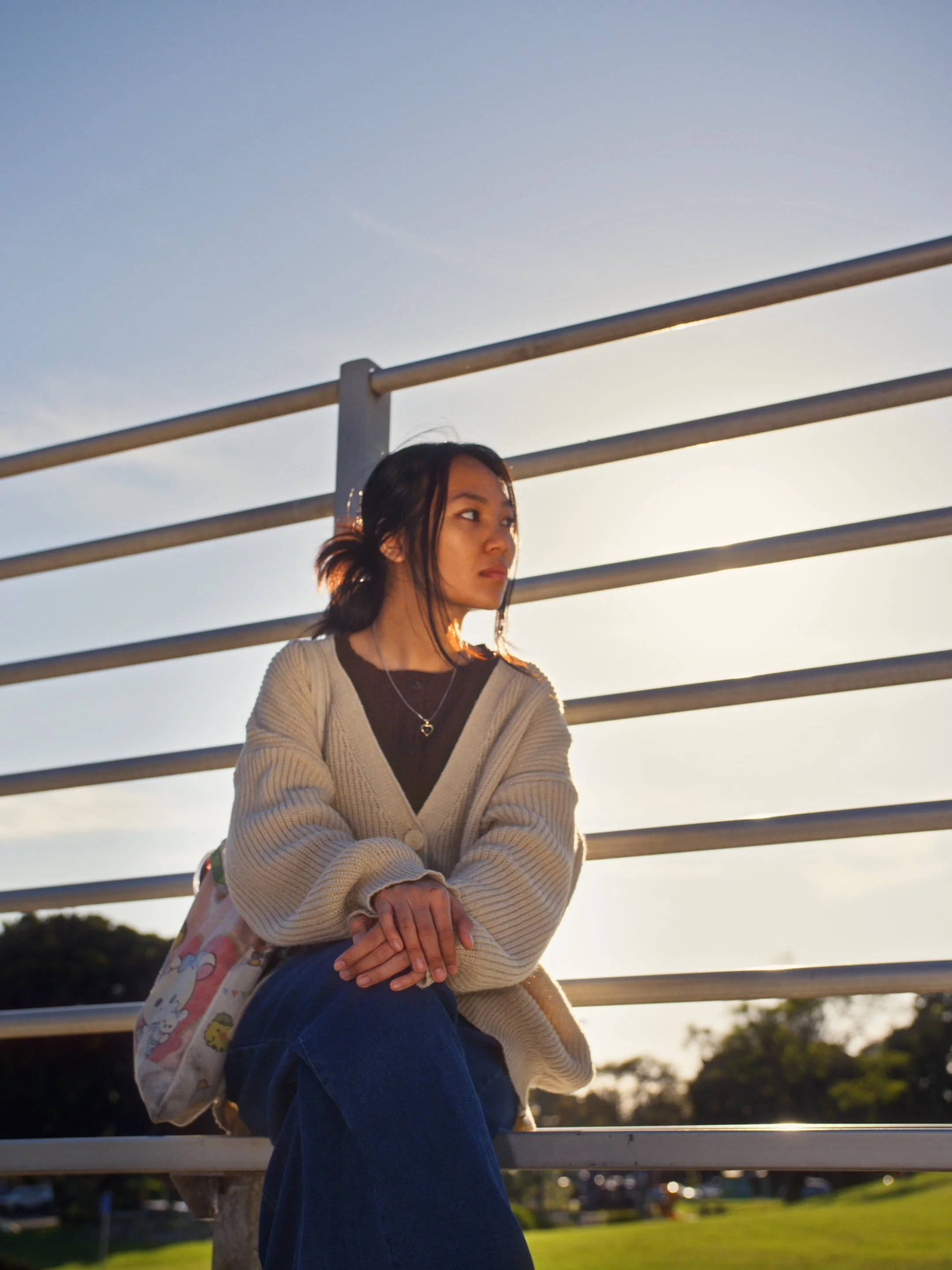 A young woman sitting outdoors on a bench with metal railings behind her, illuminated by sunlight, wearing a beige cardigan, dark top, and jeans, with trees and a grassy area in the background.