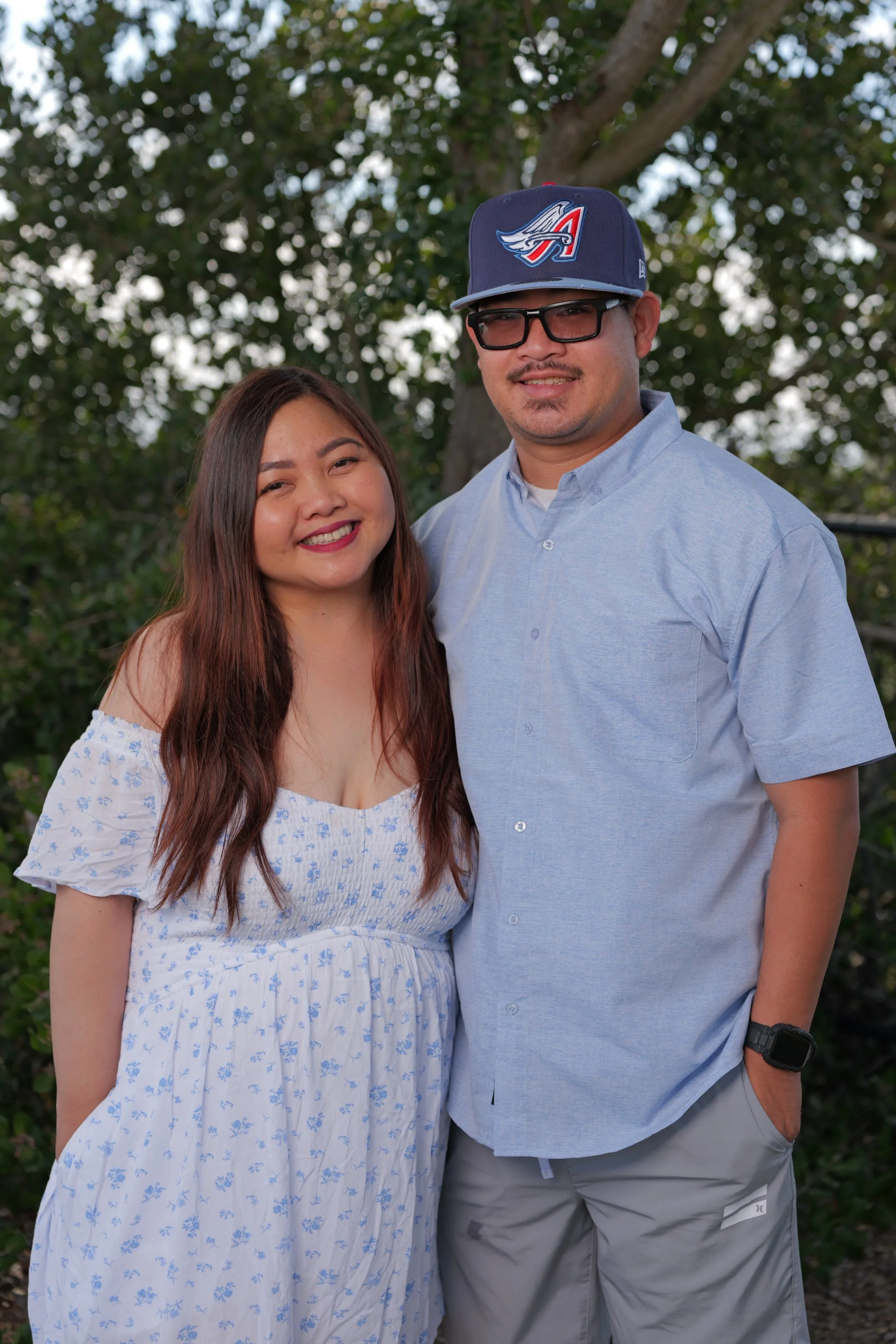 A smiling young woman with long brown hair and a man wearing glasses and a baseball cap with an 'A' logo, standing outdoors near a tree.