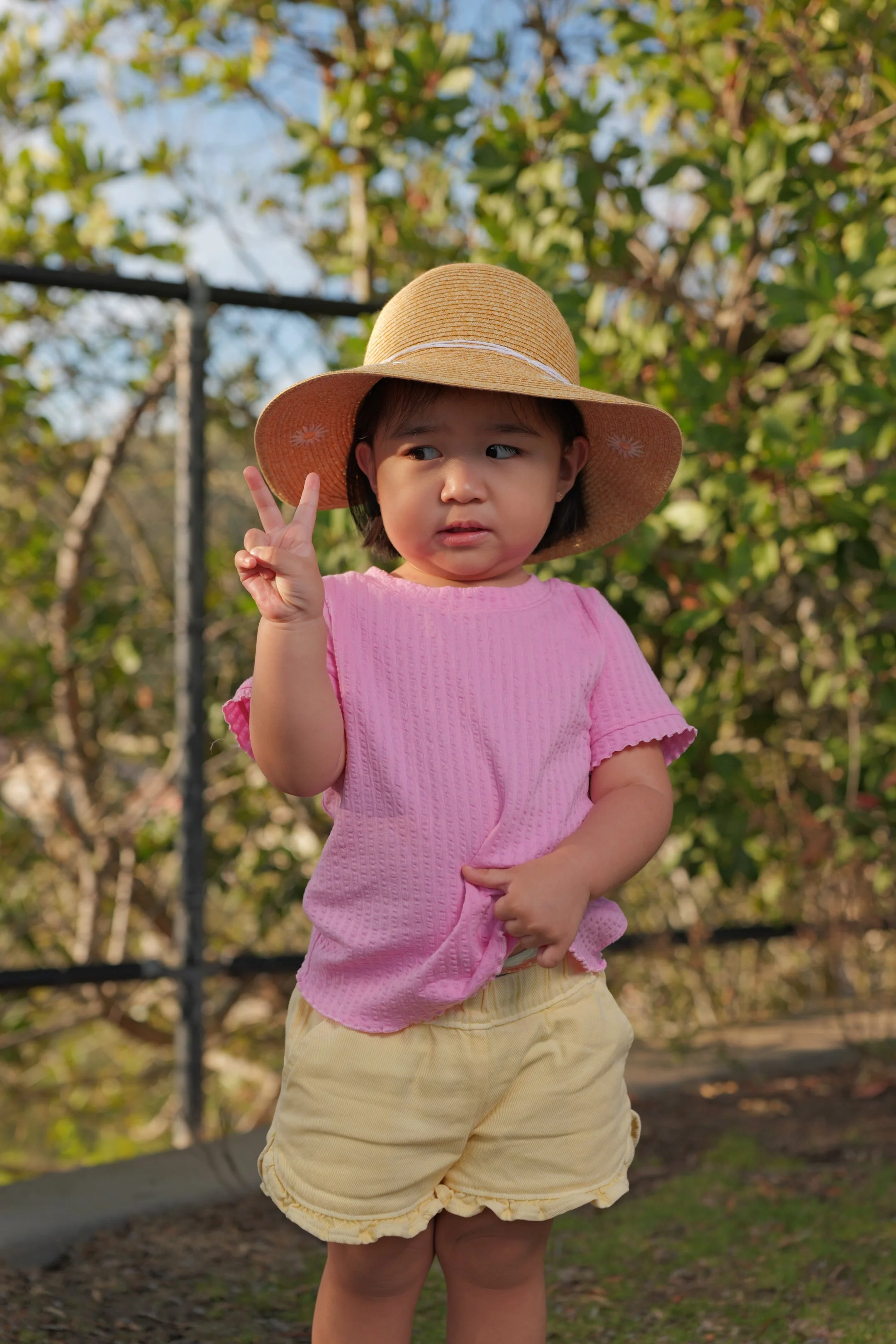 A young girl wearing a large straw sunhat, pink shirt, and yellow shorts making a peace sign gesture outdoors with greenery and trees in the background.