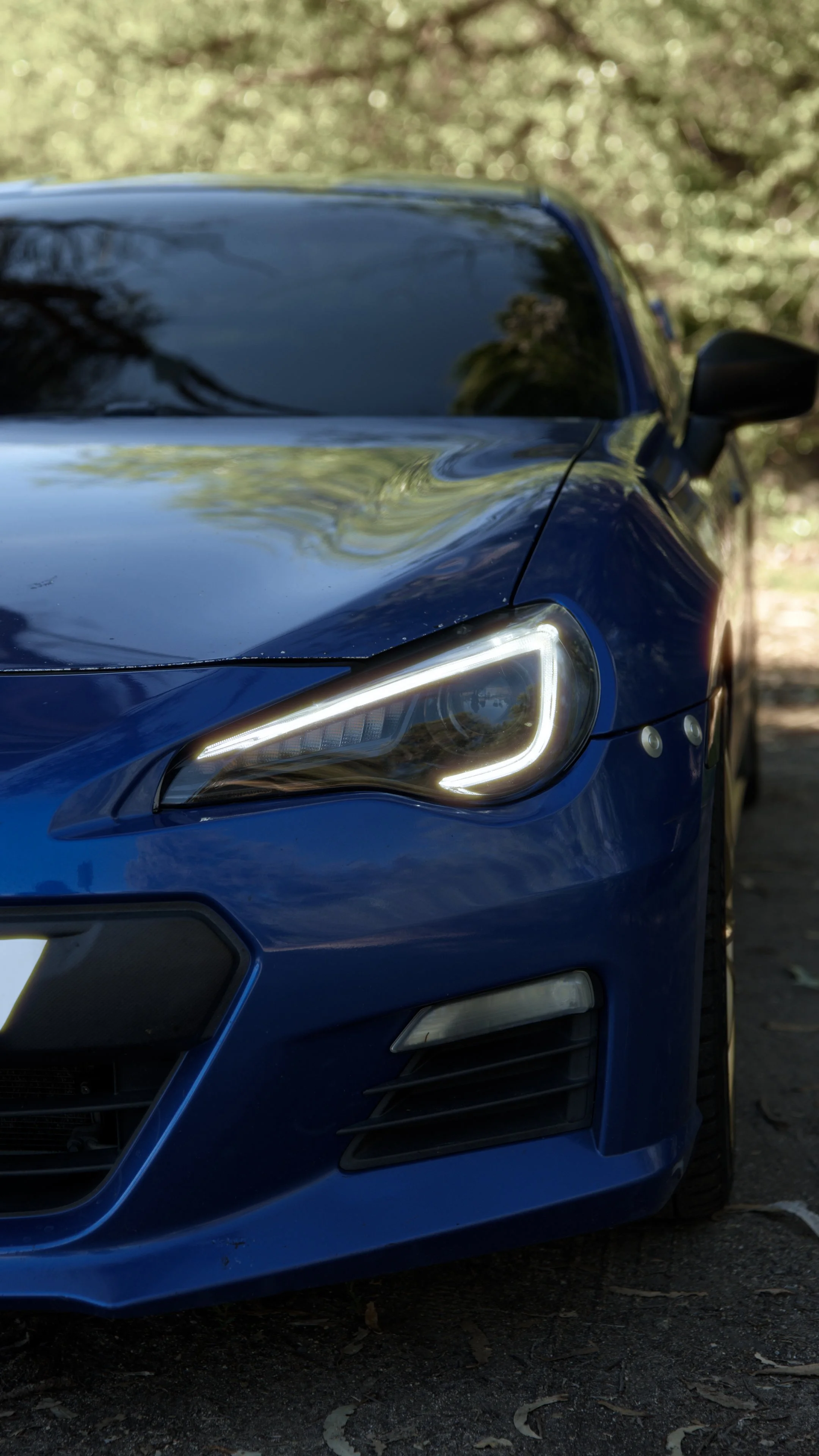 Close-up of a blue sports car with illuminated headlights parked outdoors on a dirt surface, with trees in the background.