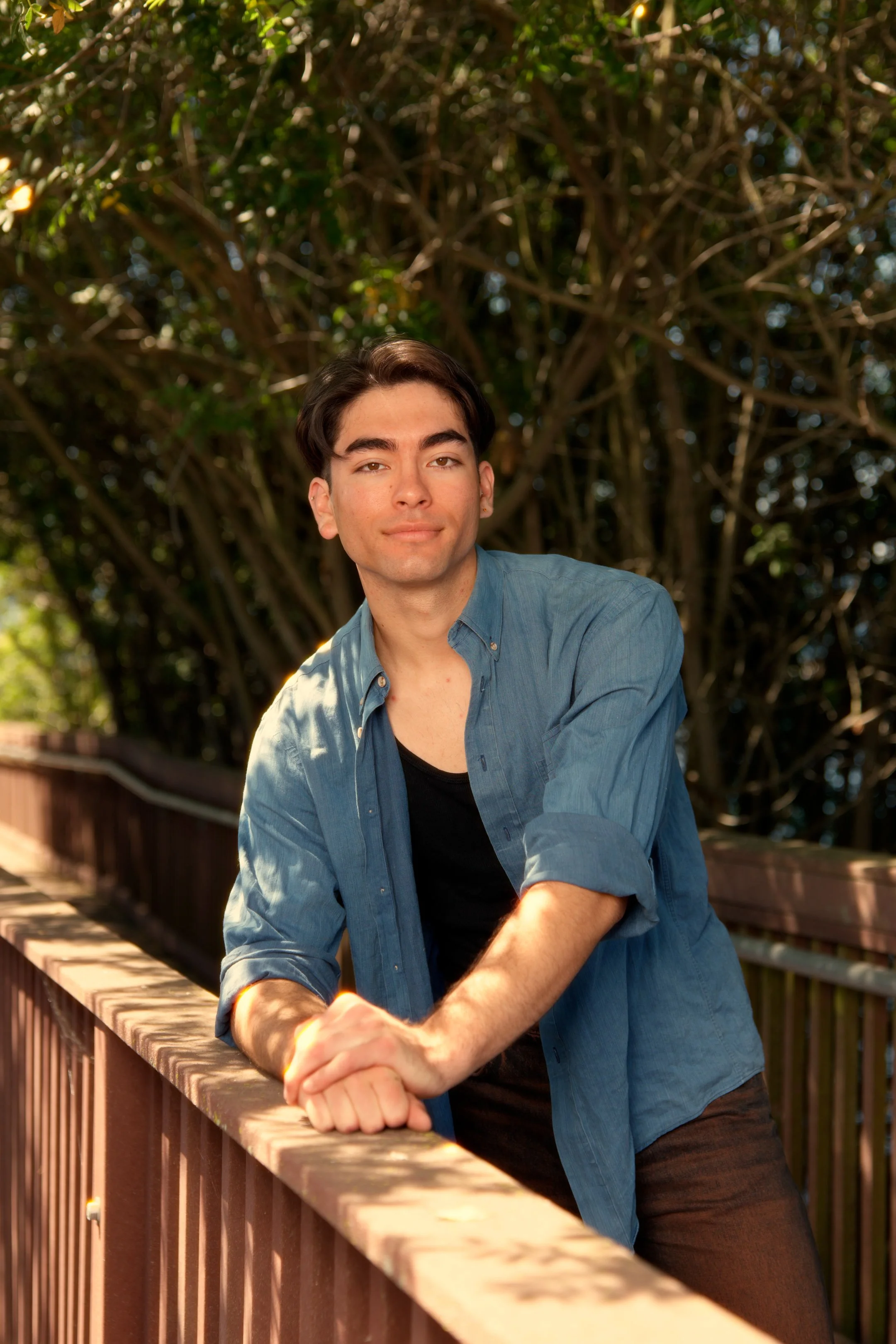 A young man with dark hair and light skin leaning on a wooden railing outdoors, surrounded by green trees and sunlight.