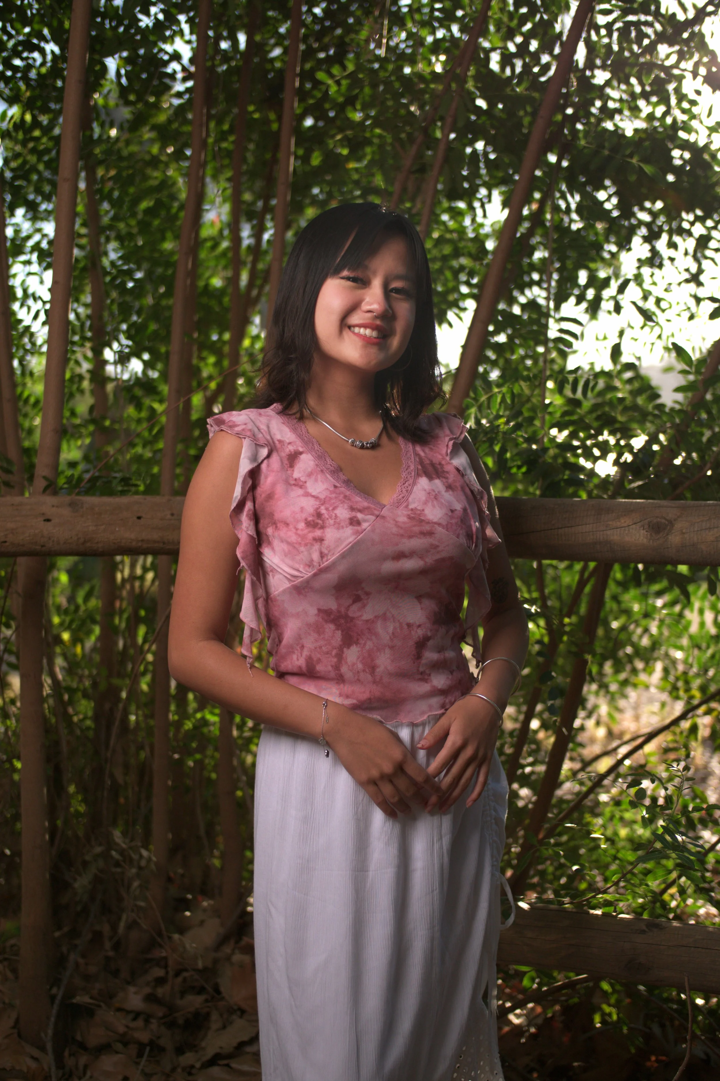 Young woman smiling outdoors in front of greenery, wearing a pink tie-dye top and white pants.