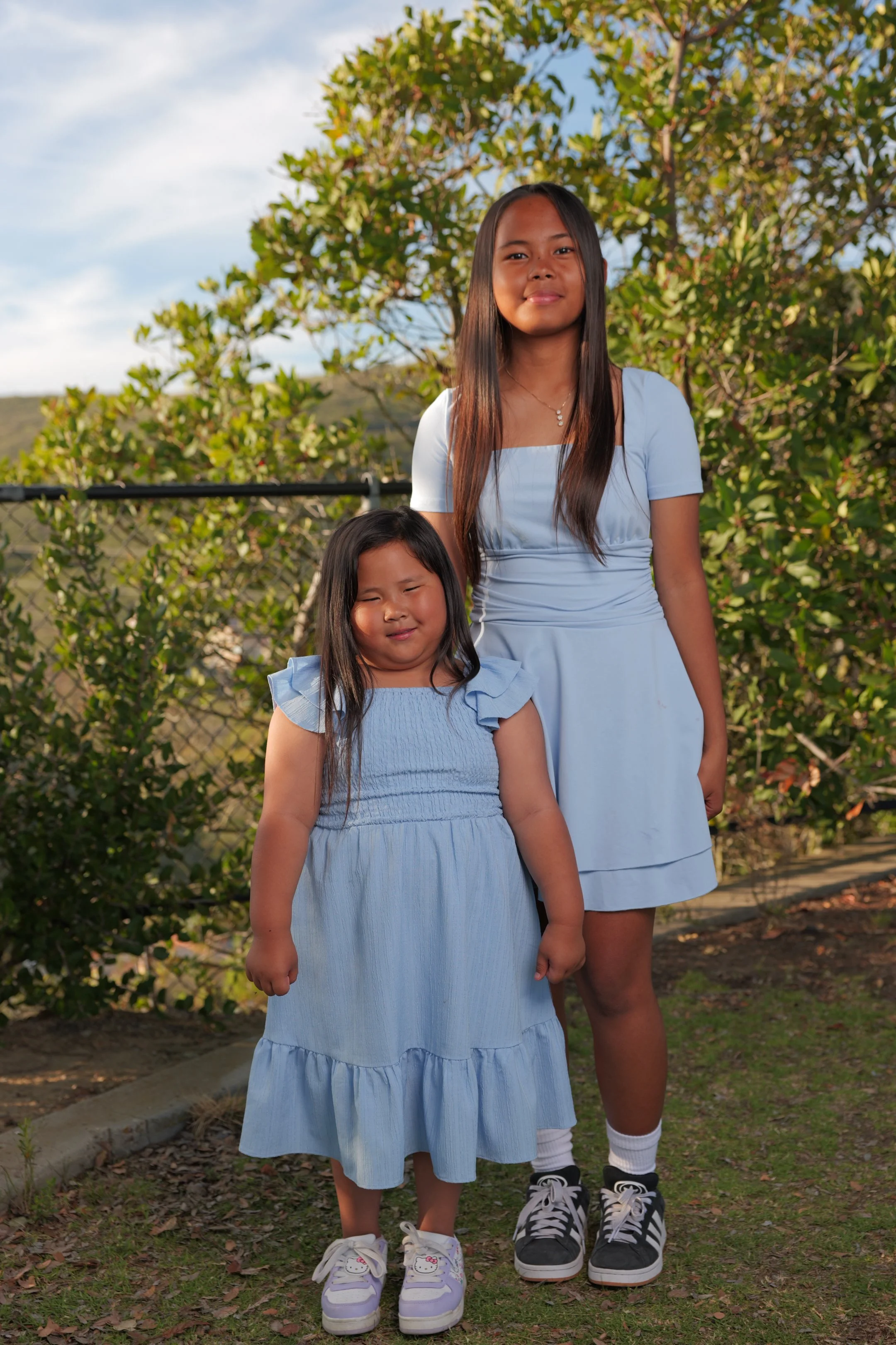 Two young girls standing outdoors in front of greenery and a chain-link fence, both wearing light blue dresses, smiling at the camera.